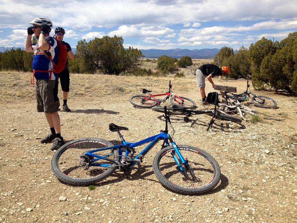 Mountain bikers taking a break on a gravel trail, with two cyclists adjusting their gear and several bikes lying on the ground. The landscape features sparse vegetation and mountains in the background under a partly cloudy sky. South Shore Lake Pueblo mountain bike trail.