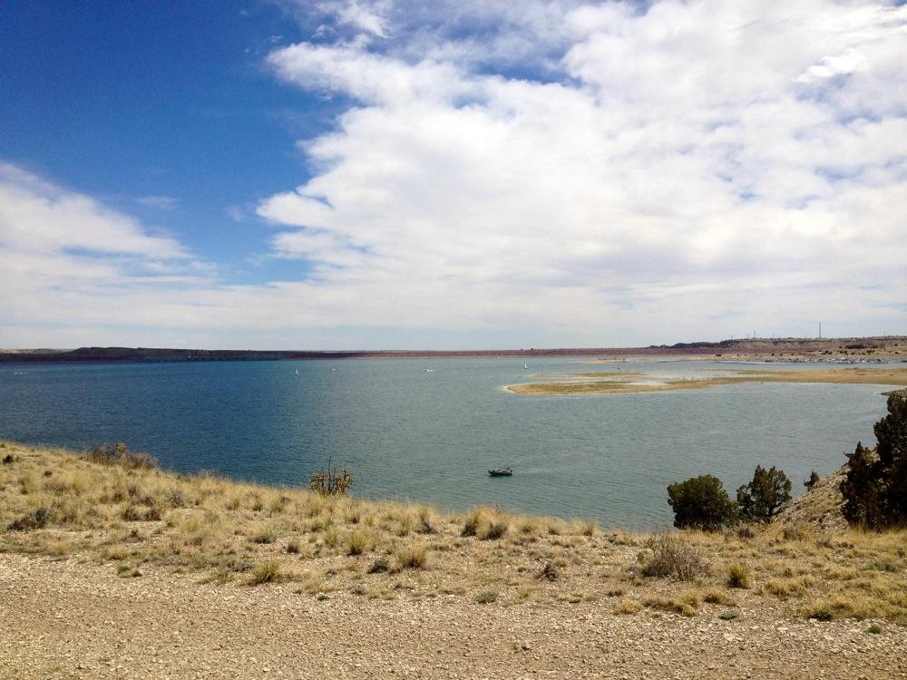 A scenic view of a tranquil lake surrounded by rolling hills and sparse vegetation under a partly cloudy sky. A small boat is visible on the water, and areas of grassland can be seen along the shore. South Shore Lake Pueblo mountain bike trail.