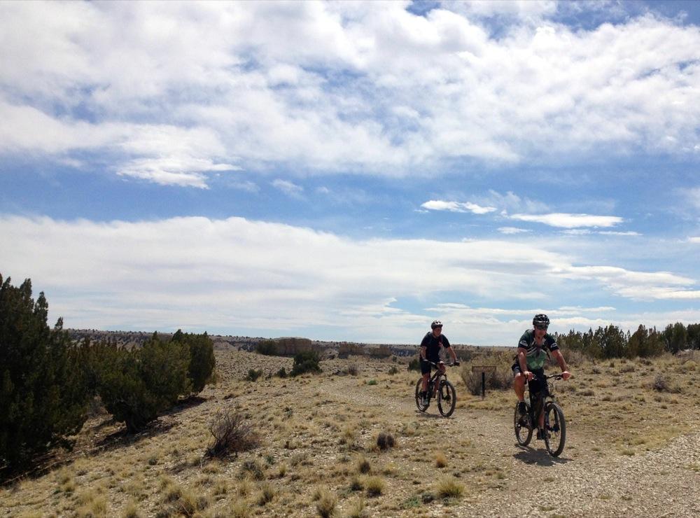 Two cyclists riding mountain bikes along a gravel trail in a wide, open landscape featuring sparse vegetation and a blue sky with scattered clouds. South Shore Lake Pueblo mountain bike trail.