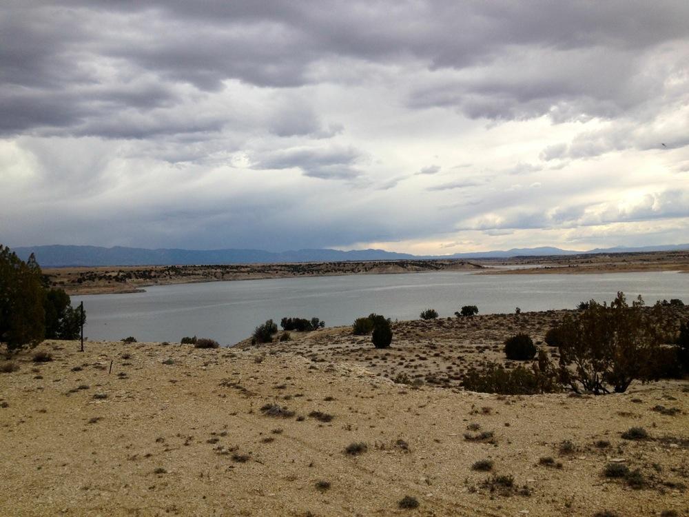 A scenic view of a calm lake surrounded by arid land, with distant mountains and a cloudy sky overhead. The shoreline is dotted with sparse vegetation and rocky terrain, creating a tranquil and natural atmosphere. North Shore Lake Pueblo State Park mountain bike trail.