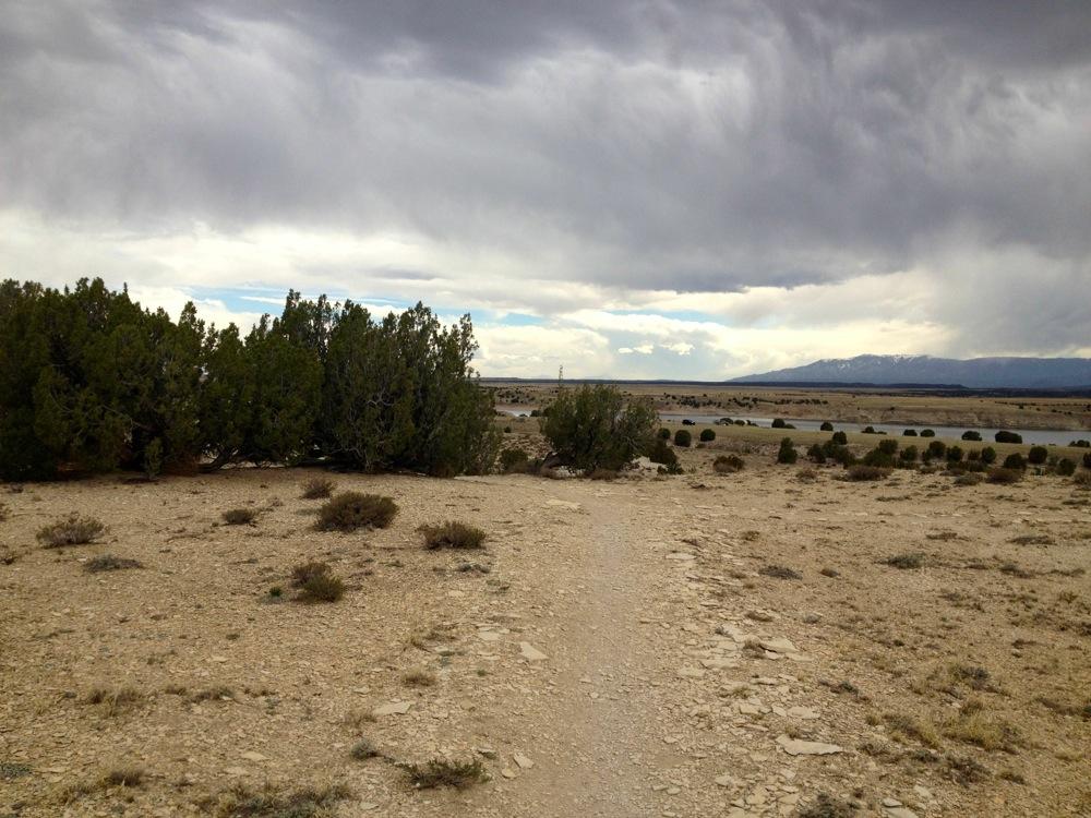 A rugged, desert landscape featuring a dirt path winding through sparse vegetation and low shrubs. The sky is overcast with varying shades of gray and hints of blue peeking through, suggesting an impending change in weather. In the distance, low hills and mountains are visible, framed by the open terrain. North Shore Lake Pueblo State Park mountain bike trail.