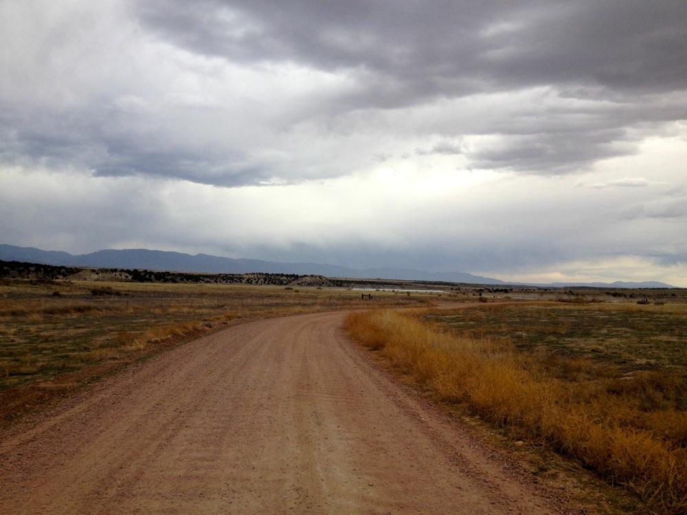 A winding dirt road extends through an open landscape under a cloudy sky, with distant mountains on the horizon and patches of dry grass on either side. North Shore Lake Pueblo State Park mountain bike trail.
