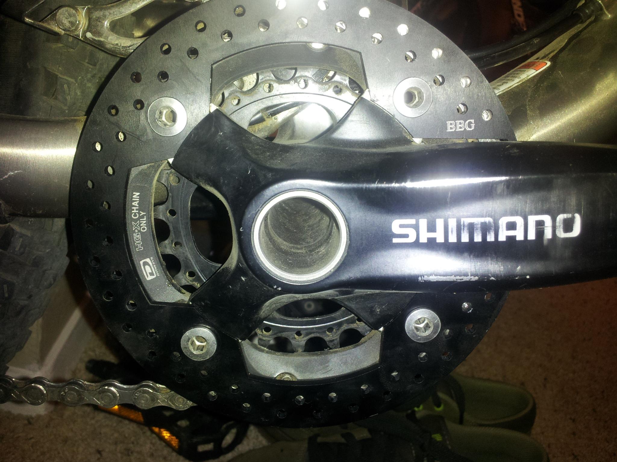 BBG Bashguard: Close-up image of a Shimano bicycle crankset with a black chainring, showing details such as bolt holes and branding. The background includes parts of a bike tire and footwear.