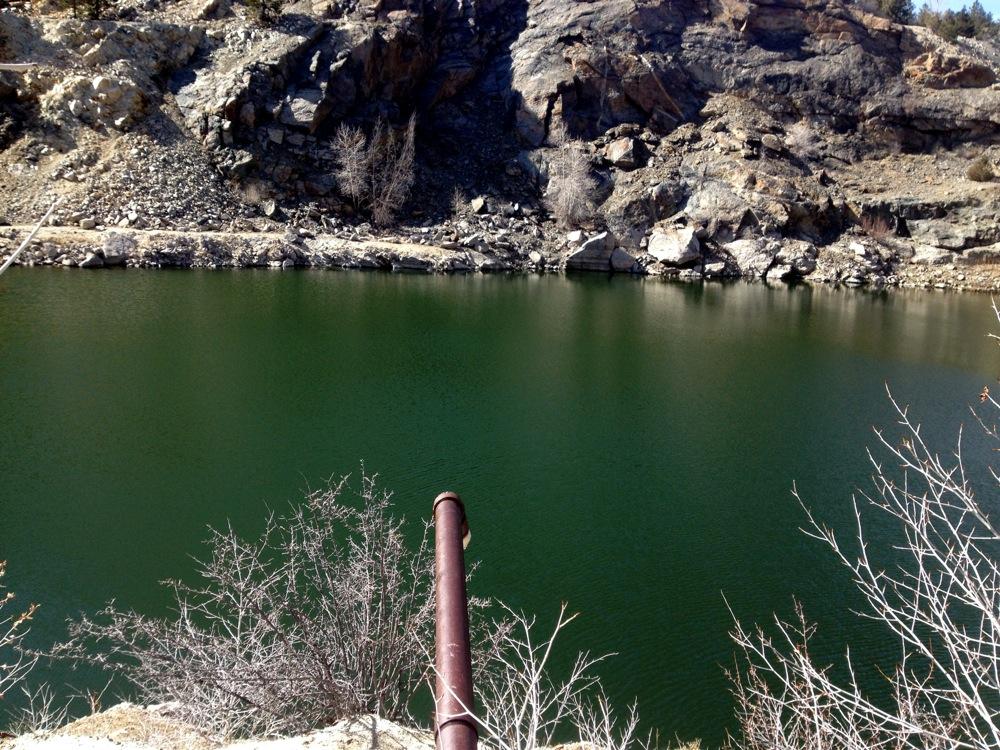 A tranquil green lake surrounded by rocky cliffs, with a rusty pipe extending toward the water's edge. Sparse vegetation, including some bare bushes, is visible in the foreground. The scene is set in a natural landscape, suggesting a remote or rugged environment. Aspen Ridge / Road #185 mountain bike trail.