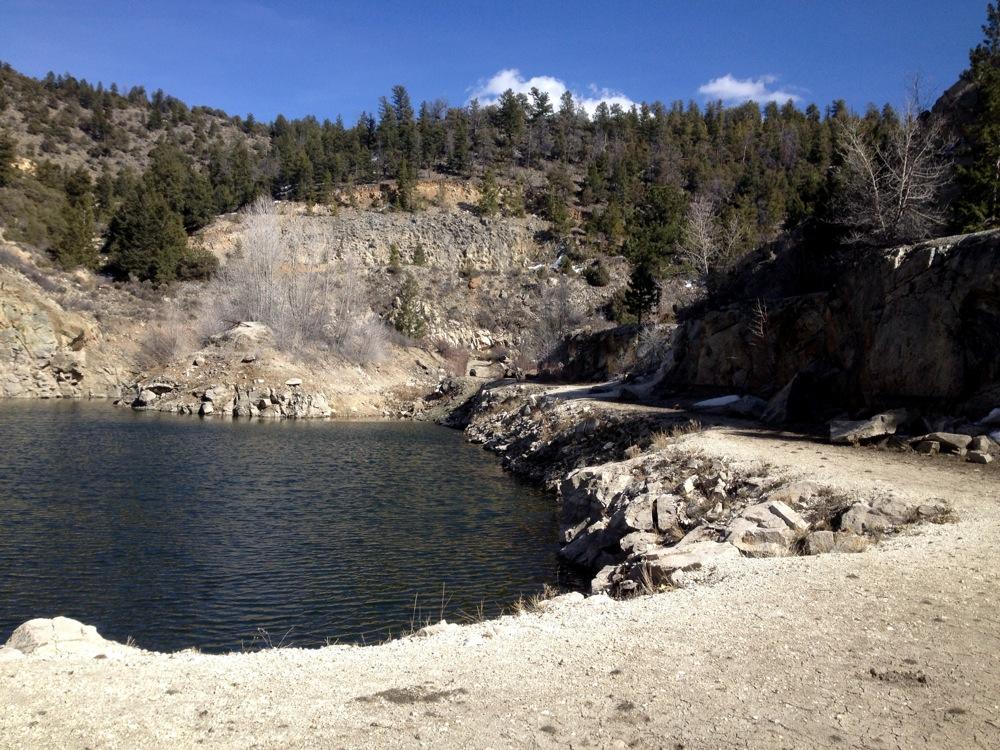 A serene landscape featuring a calm body of water bordered by rocky shoreline and a dirt path. Surrounding the water are steep, rocky hillsides covered with sparse vegetation and trees, under a clear blue sky with a few clouds in the background. The setting suggests a remote and natural environment, ideal for outdoor activities. Aspen Ridge / Road #185 mountain bike trail.