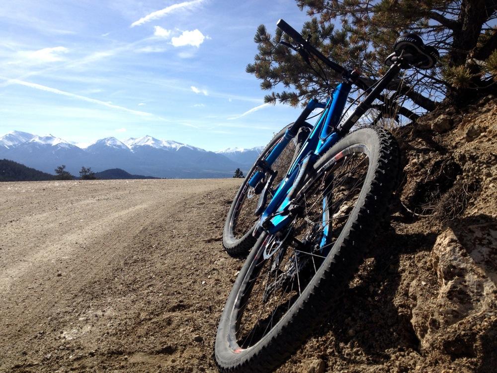 Mountain bike leaning against a rocky hillside, with a dirt road winding through a mountainous landscape in the background. Snow-capped mountains and a clear blue sky are visible in the distance. Road #184 mountain bike trail.