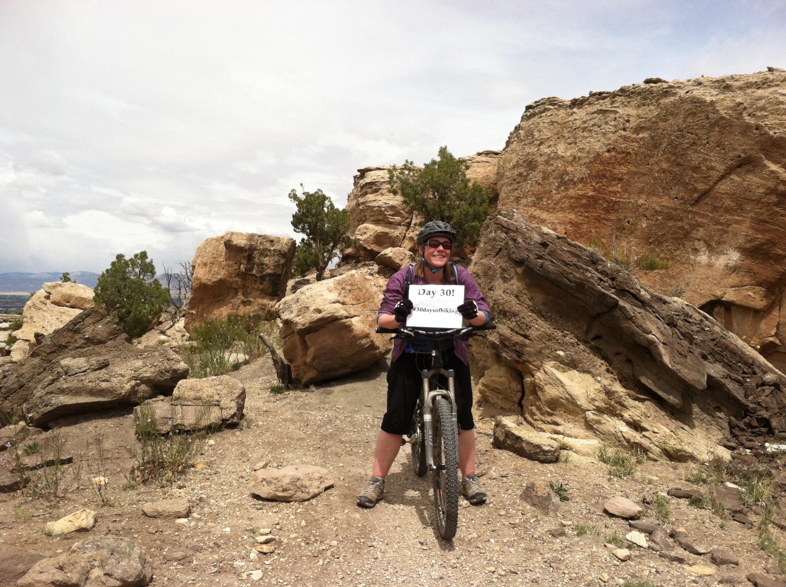 A woman wearing a helmet and cycling gear stands beside her mountain bike on a rocky trail, holding a sign that says "Day 30!" The background features large rocks and patches of greenery under a cloudy sky. Lunch Loops mountain bike trail.