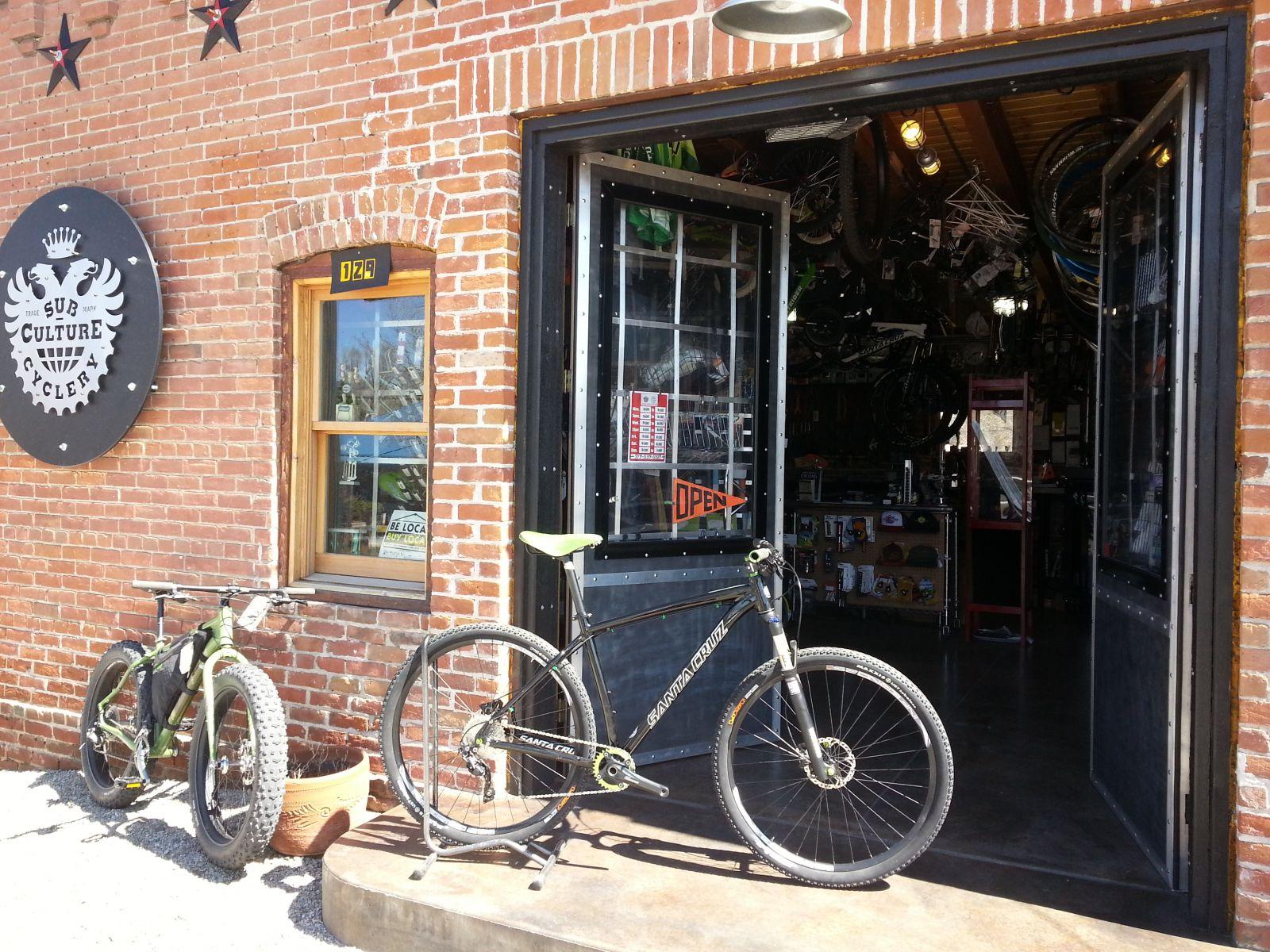 Alt text: Entrance of a bike shop featuring a brick façade, with two bicycles parked outside—one fat bike and one mountain bike. The open door showcases a variety of bicycles and accessories inside, with a visible "OPEN" sign. A sign above the door reads "Sub Culture Cycle."