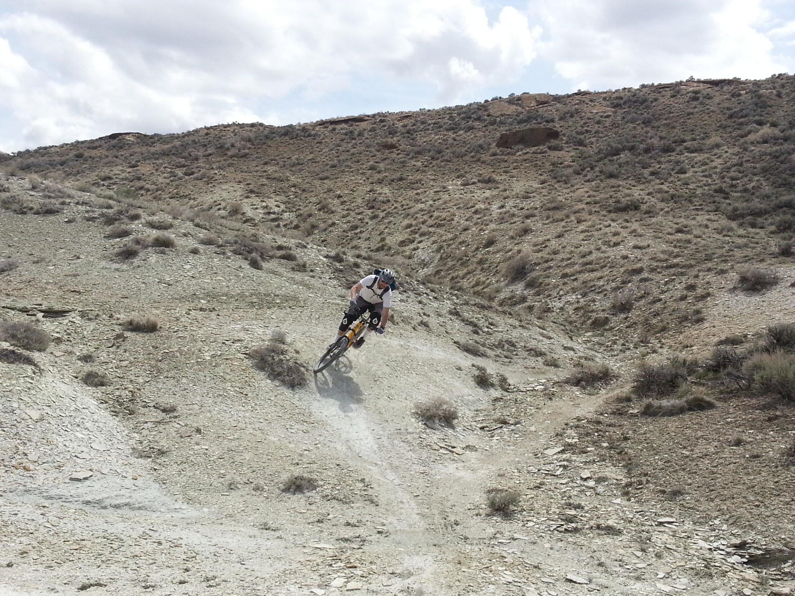 A mountain biker leaning into a turn on a rocky, uneven trail in a rugged, dry landscape under a cloudy sky. Cruiser mountain bike trail.