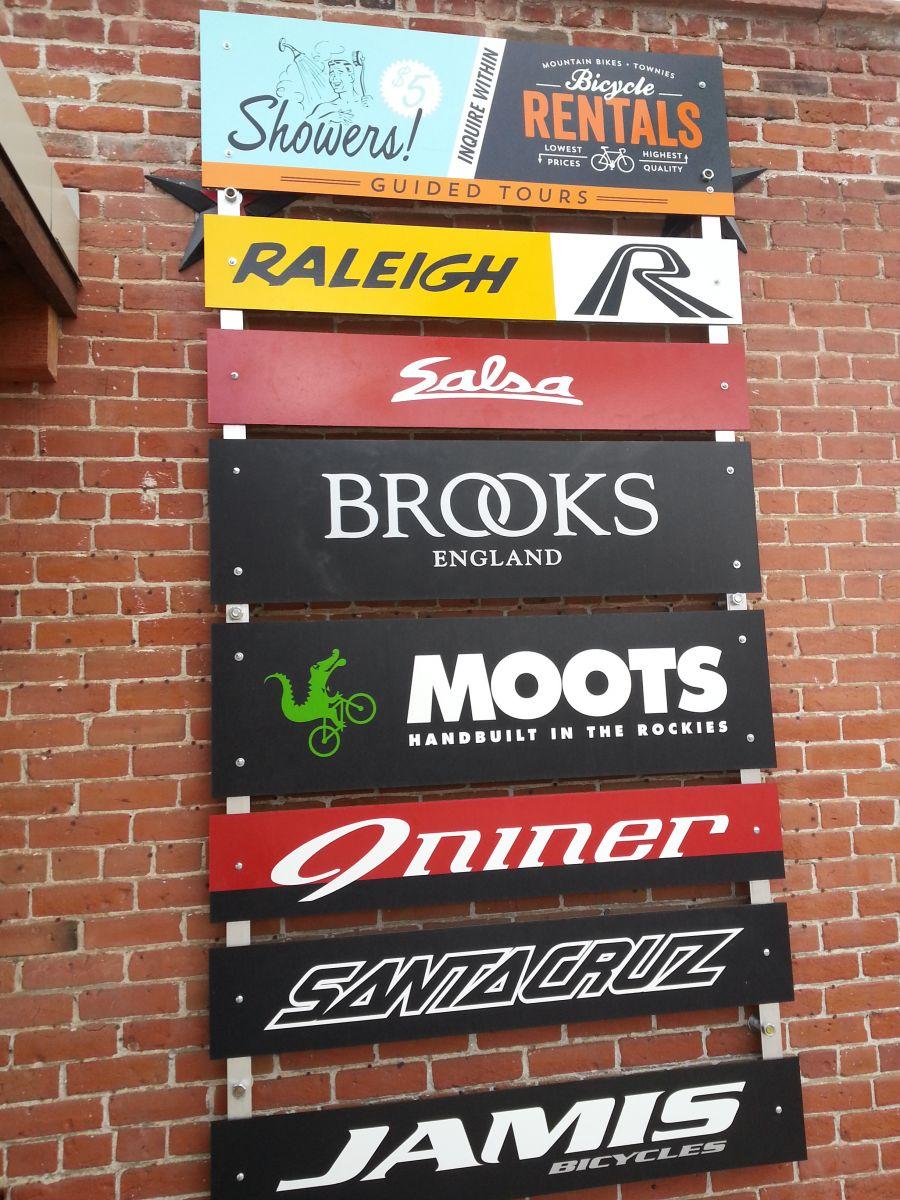 A vertical display of signs mounted on a brick wall, featuring various bicycle brands and services. The list includes "Showers!" at the top for $5, followed by "Bicycle Rentals," "Guided Tours," "Raleigh," "Salsa," "Brooks England," "Moots," "Oniner," "Santa Cruz," and "Jamis Bicycles." Each sign has a distinct color and design, highlighting the brand names.