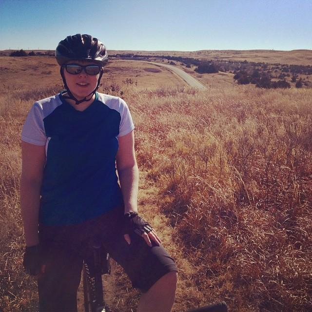 Person in cycling gear, wearing a helmet and sunglasses, seated on a bicycle in a grassy area with a clear blue sky. In the background, a winding road and rolling hills are visible. Switchgrass mountain bike trail.