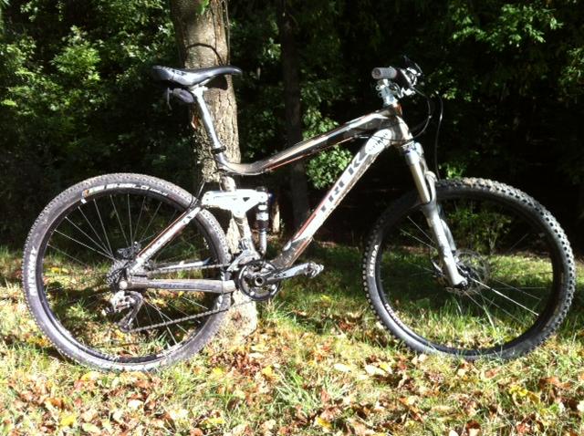 Trek Fuel EX 9: A mountain bike leaning against a tree, surrounded by grass and fallen leaves. The bike features a black and silver frame, with thick tires designed for off-road riding. Sunlight filtering through the trees highlights the bike's frame and parts.