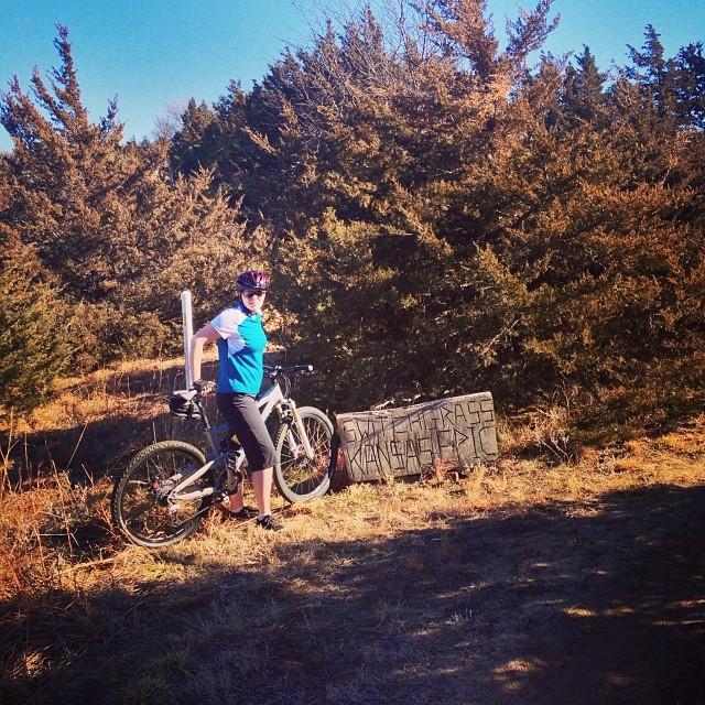 A person wearing a blue shirt and black shorts stands next to a bicycle, leaning against it in a grassy area surrounded by shrubs. A sign nearby indicates the location. The scene is outdoors with clear blue skies in the background. Switchgrass mountain bike trail.