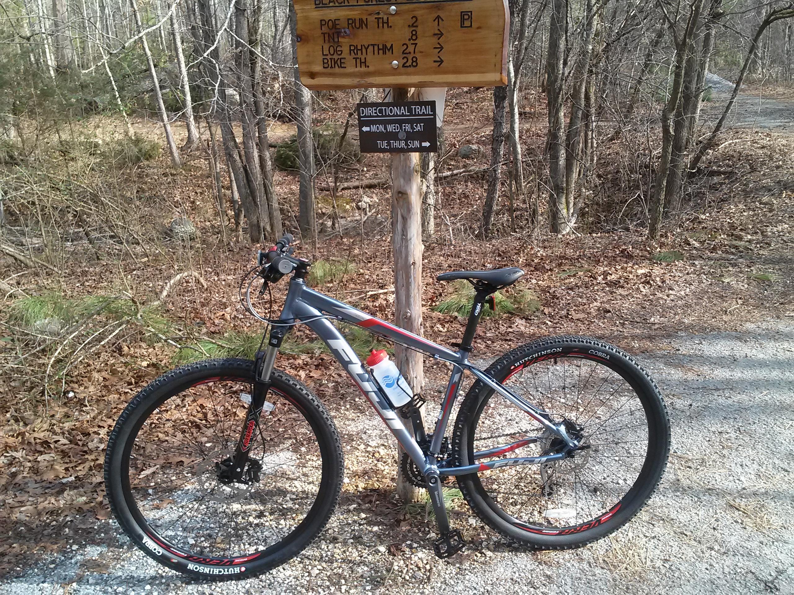 Fuji nevada 1.5 29er: A mountain bike is positioned beside a trail sign in a wooded area. The sign indicates the names and distances of various trails, including "Poe Run TH," "Log Rhythm," and "Bike TH." The background features trees and fallen leaves, suggesting an outdoor recreational setting. A water bottle is attached to the bike's frame.
