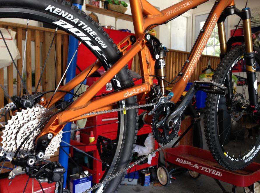 Turner Burner: Close-up of an orange mountain bike with prominent components visible, including the rear wheel, cassette, and chain. The bike is positioned in a garage, showcasing various tools and a red toolbox in the background.