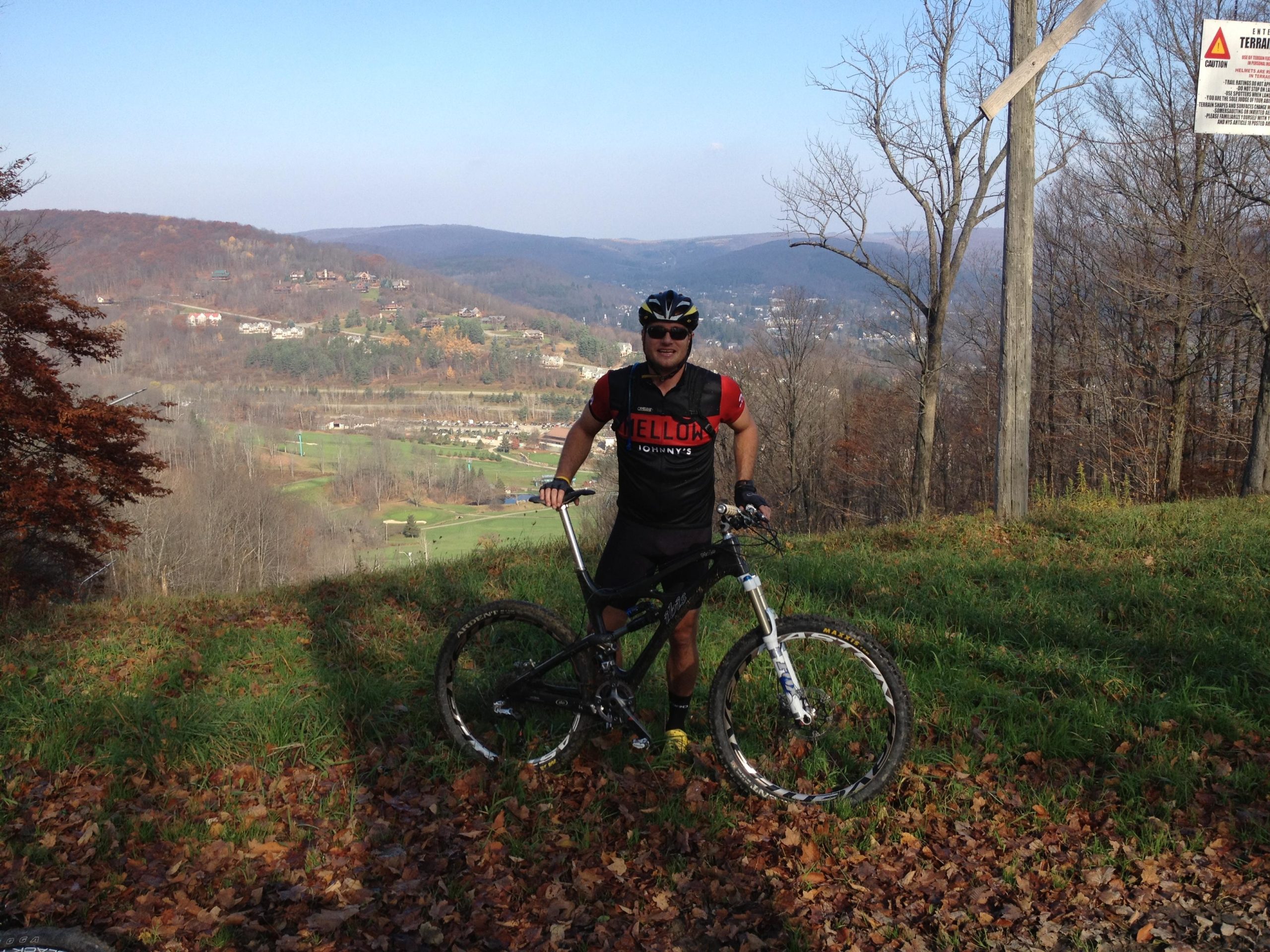A person in a cycling outfit stands next to a mountain bike on a grassy hillside, overlooking a scenic valley with autumn foliage. The background features distant hills and a clear blue sky. A caution sign is visible nearby, indicating a warning about the terrain. Ellicottville Epic mountain bike trail.