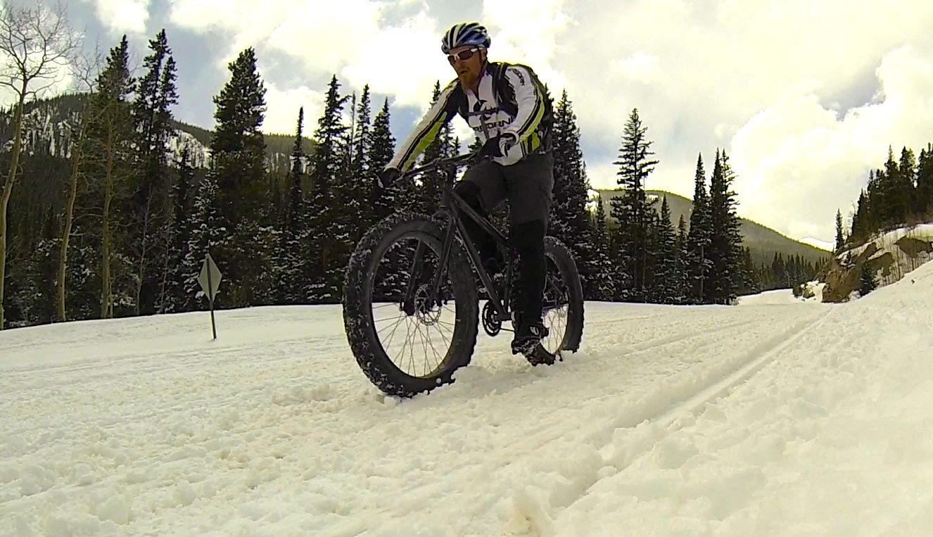 A cyclist rides a fat bike on a snowy trail surrounded by tall evergreen trees and a mountain landscape, with a partly cloudy sky overhead. Cottonwood Pass Road mountain bike trail.