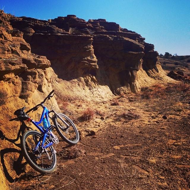 A blue mountain bike leaning against a rocky cliff under a clear blue sky, with dry grass and dirt in the foreground. Switchgrass mountain bike trail.