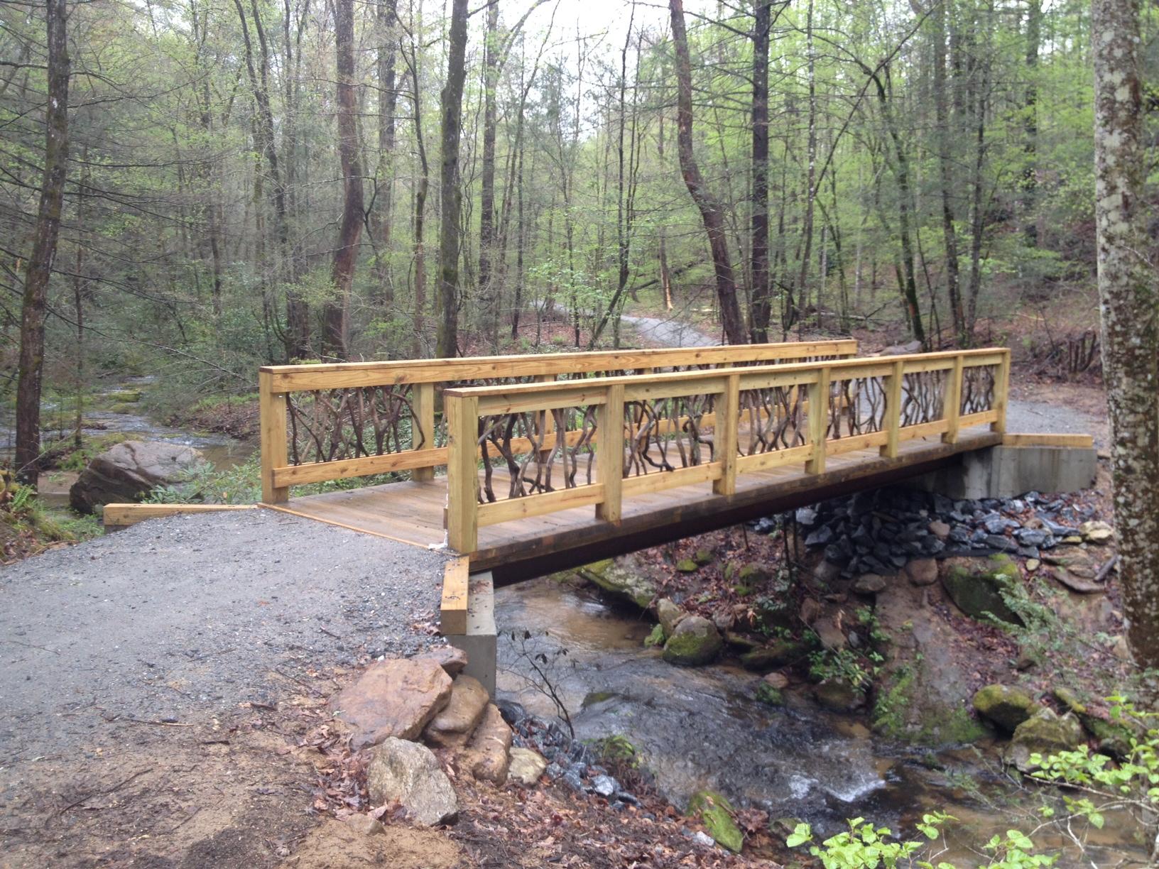 A wooden bridge spans a small stream in a lush, green forest. The bridge features a decorative railing made of branches, and the surrounding area is filled with trees and undergrowth. A gravel path leads up to the bridge, while the gentle flow of water can be seen beneath it. Buffalo Creek Park: The Head, The Heart &amp; The Tail (HHT) mountain bike trail.