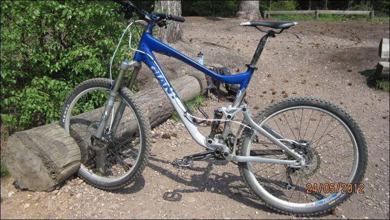 Giant Trance: A blue and silver mountain bike parked next to a log on a gravel path surrounded by greenery. The bike features thick tires and a sturdy frame, indicating it's designed for off-road cycling. Sunlight filters through the trees in the background, and a water bottle can be seen on the ground nearby.