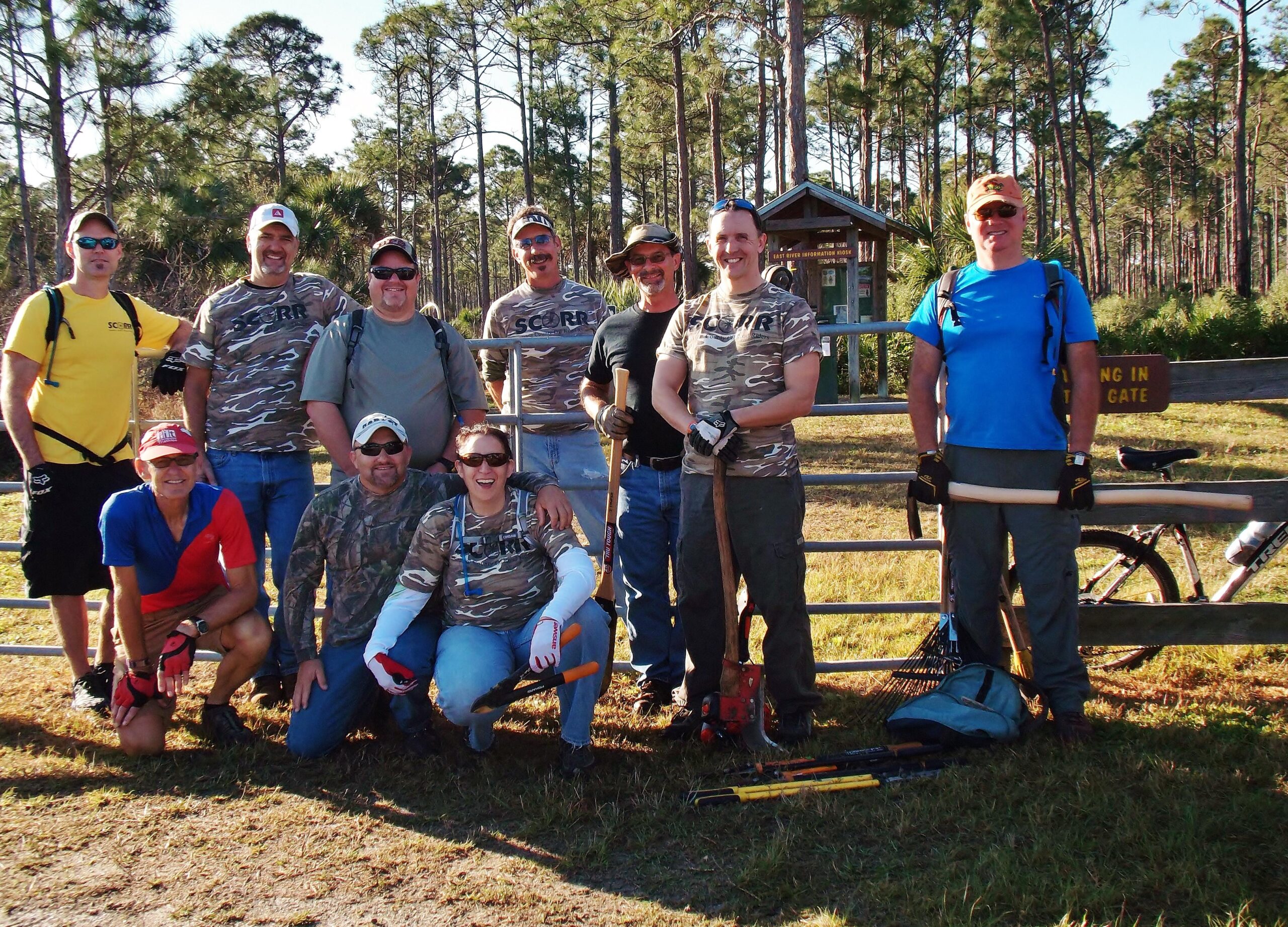 A group of ten volunteers poses for a photo while standing in a park. They are wearing matching camouflage shirts and casual attire, with some holding gardening tools and equipment. The background features tall pine trees and a park sign. The group appears cheerful and ready for outdoor activities. Palmetto Trail mountain bike trail.