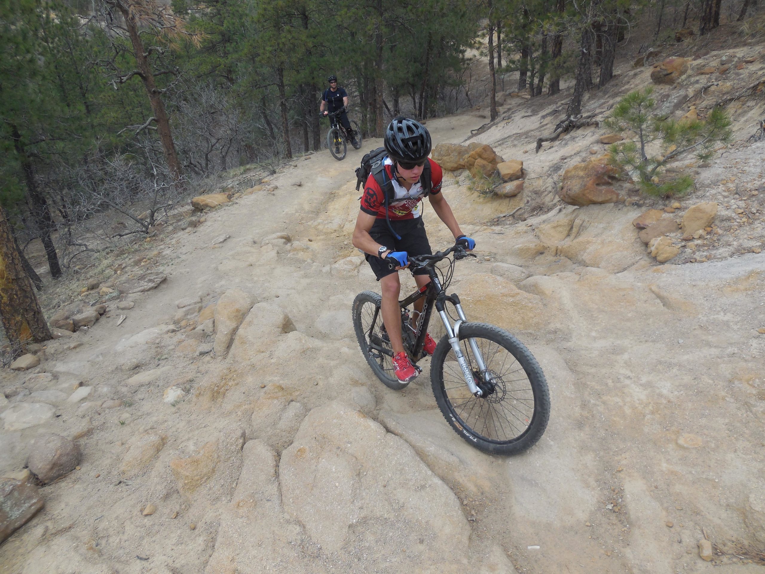 A mountain biker navigates a rocky trail in a wooded area, with trees in the background. Another cyclist is visible in the distance, also riding along the path. The terrain appears challenging, with loose stones and a steep incline. Palmer Park mountain bike trail.