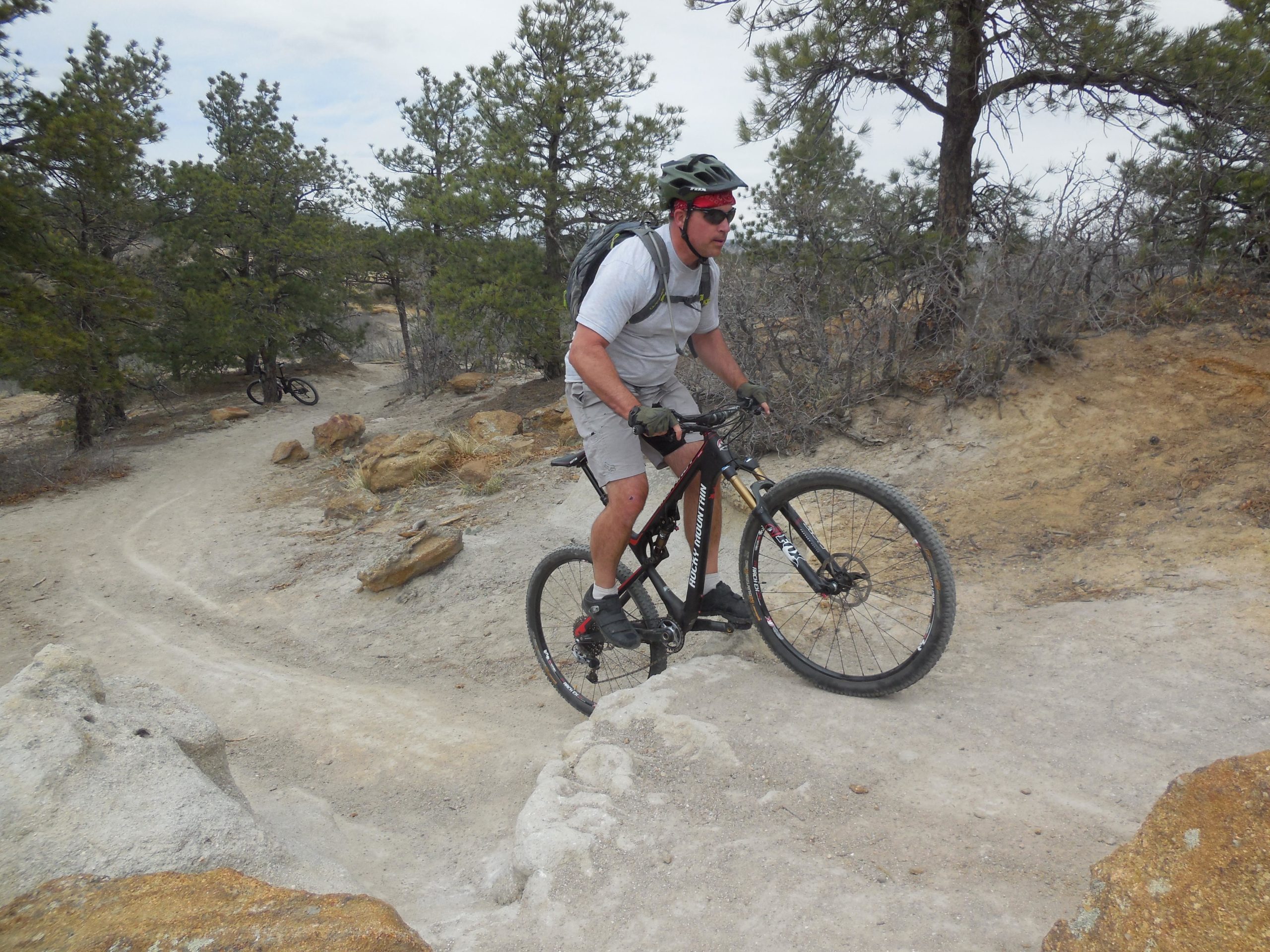 A person riding a mountain bike on a dirt trail surrounded by trees and rocky terrain. The cyclist is wearing a helmet and sports gear, navigating a curved path. In the background, another bike is partially visible among the trees. The atmosphere suggests an outdoor wilderness adventure. Palmer Park mountain bike trail.