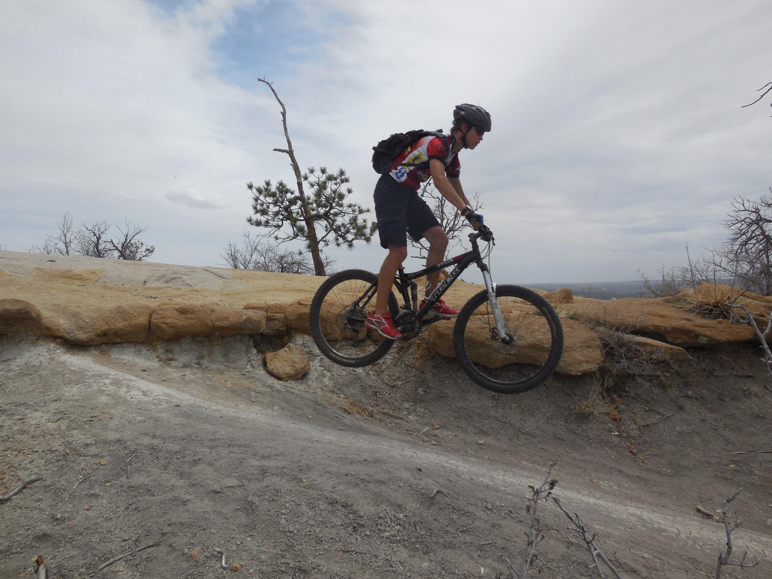 A mountain biker in mid-air, jumping off a rock formation on a dirt trail. The rider wears a helmet and sports gear, with a backpack and red biking shoes. The landscape features scattered trees and rugged terrain under a cloudy sky. Palmer Park mountain bike trail.