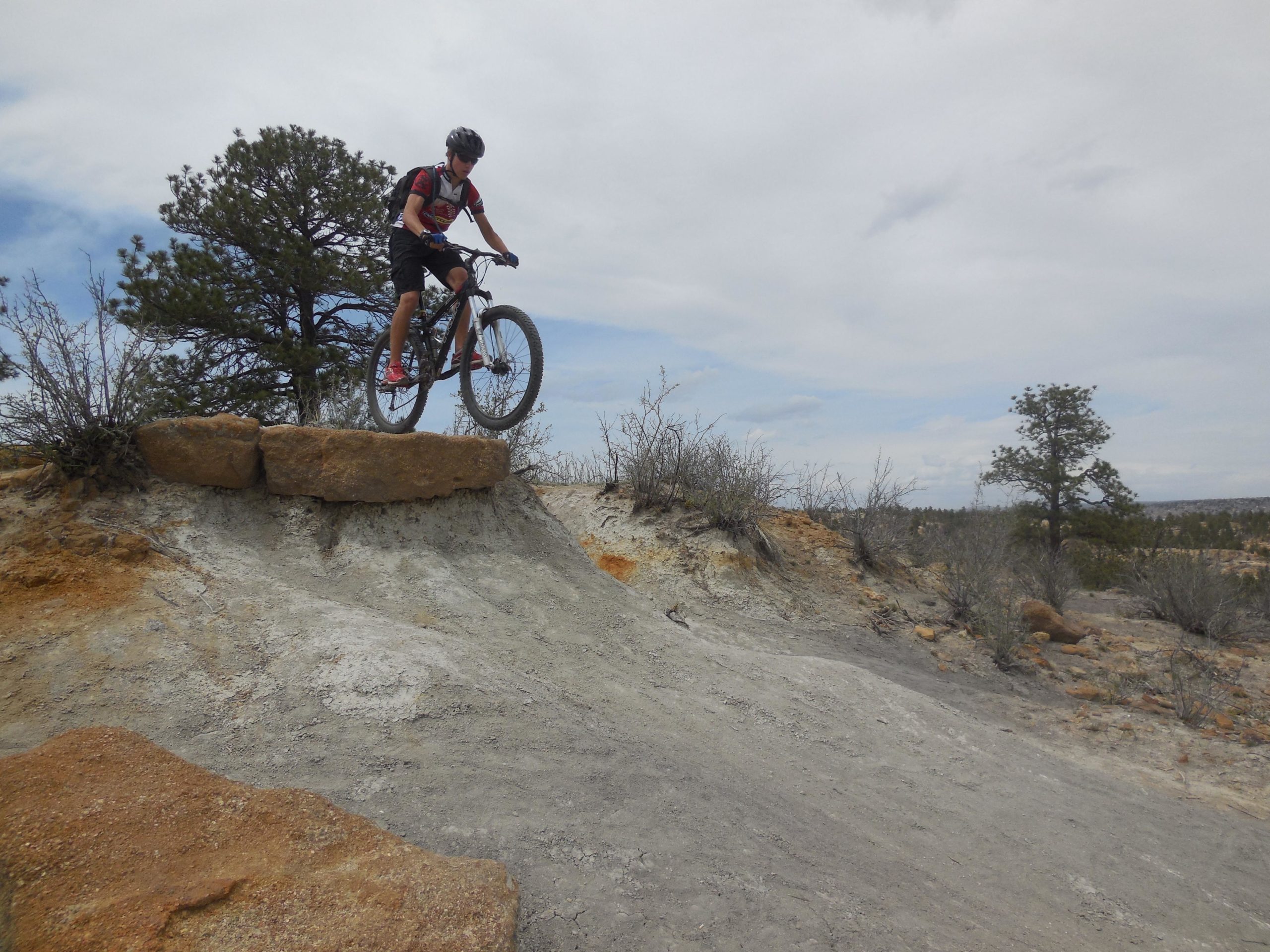 A mountain biker jumping off a rocky ledge on a dirt trail, with a sparse landscape of shrubs and trees in the background under a cloudy sky. Palmer Park mountain bike trail.