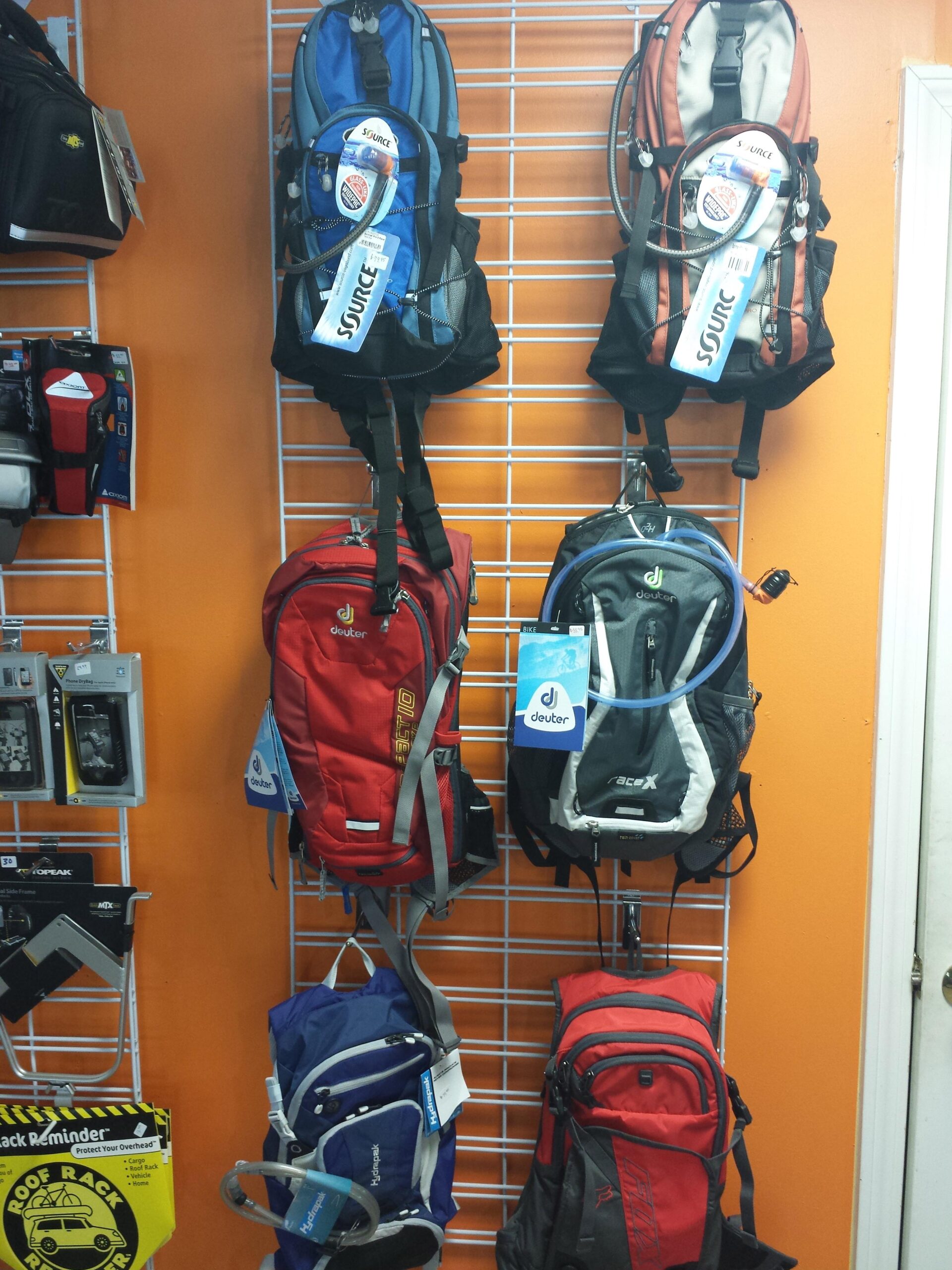 A display of various backpacks hanging on a white grid wall, featuring multiple designs and colors, including blue, red, gray, and orange. Each backpack is tagged with brand labels, showcasing different styles suitable for outdoor activities. The background is an orange wall, enhancing the visibility of the backpacks.