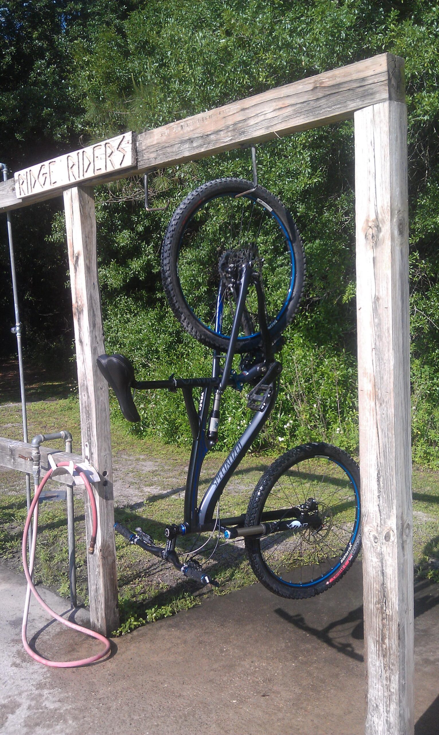 Specialized Camber Pro: A black mountain bike hanging upside down on a wooden bike rack with the words "RIDGE RIDERS" carved into the beam above. A red hose lies coiled on the ground nearby, and the backdrop features greenery in a sunny outdoor setting.