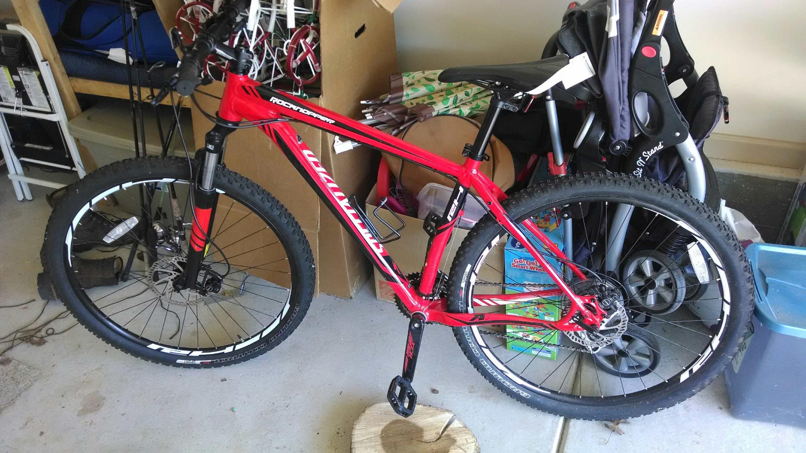 Specialized Rockhopper Expert Disc 29er: A red mountain bike resting on a wooden block in a garage, surrounded by various items including boxes, a stroller, and fishing rods. The bike features thick tires and a disc brake, emphasizing its rugged design.