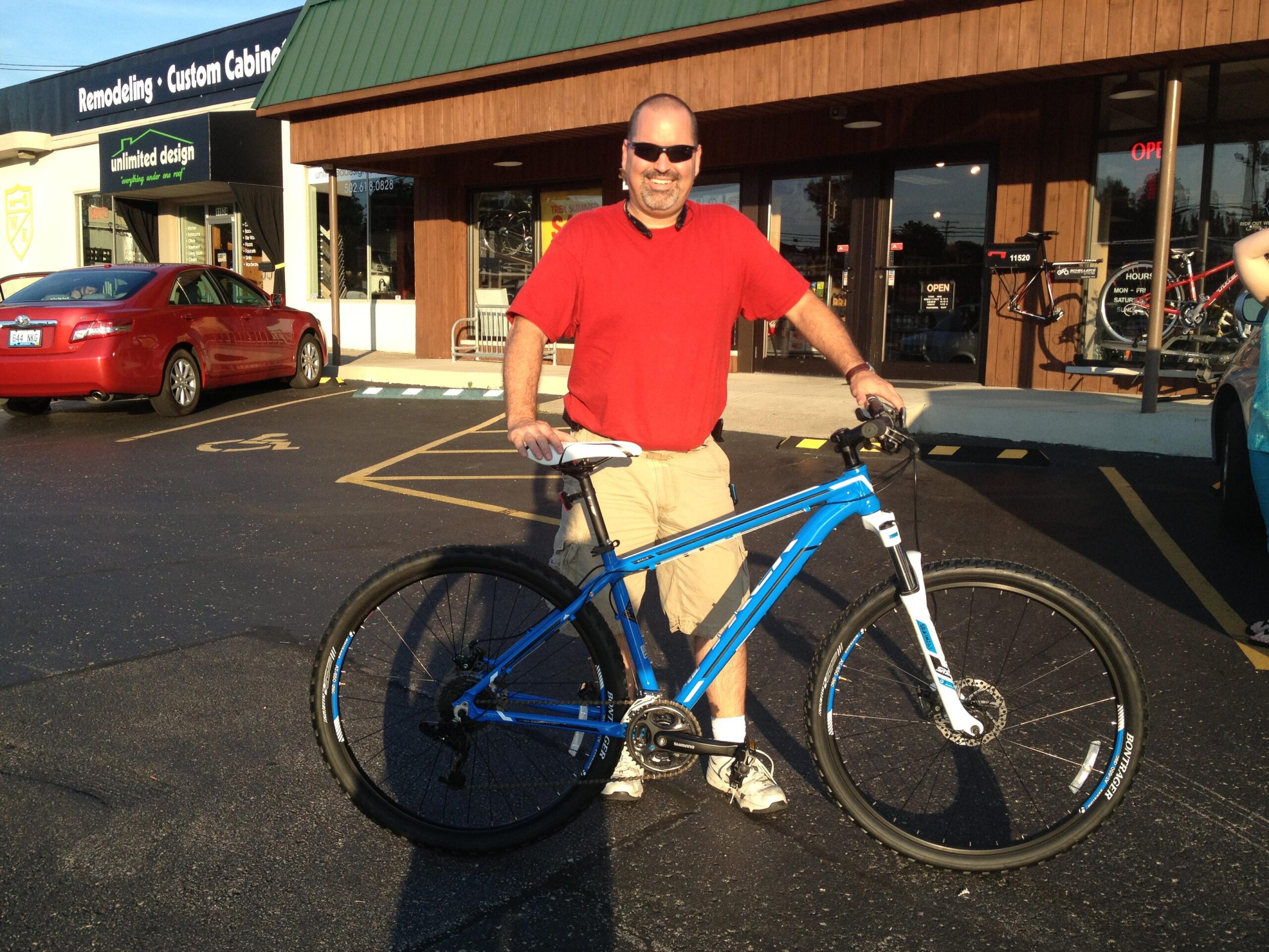 Trek Marlin: A man in a red shirt stands outside a bicycle shop, smiling and holding a blue mountain bike. The shop features a wooden facade and a green roof. A red car is parked nearby, and bicycles are displayed in the shop windows. Sunlight is shining, suggesting a clear day.