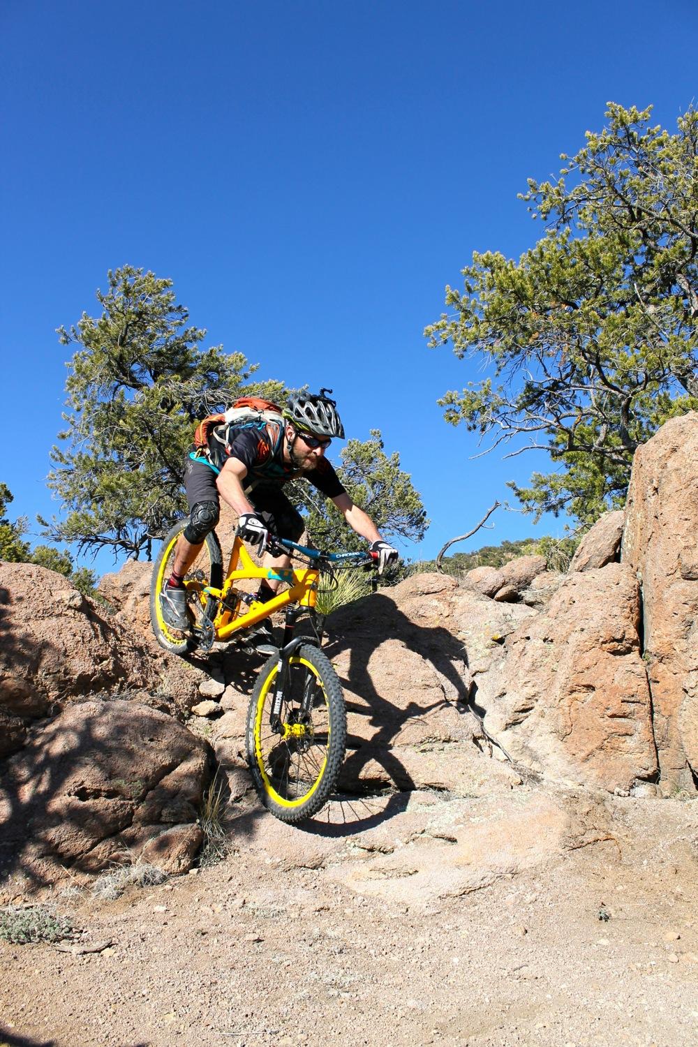 A mountain biker navigating a rocky terrain, performing a jump on a bright yellow bicycle. The rider is wearing a helmet and protective gear, with trees in the background under a clear blue sky. Unkle Nazty mountain bike trail.