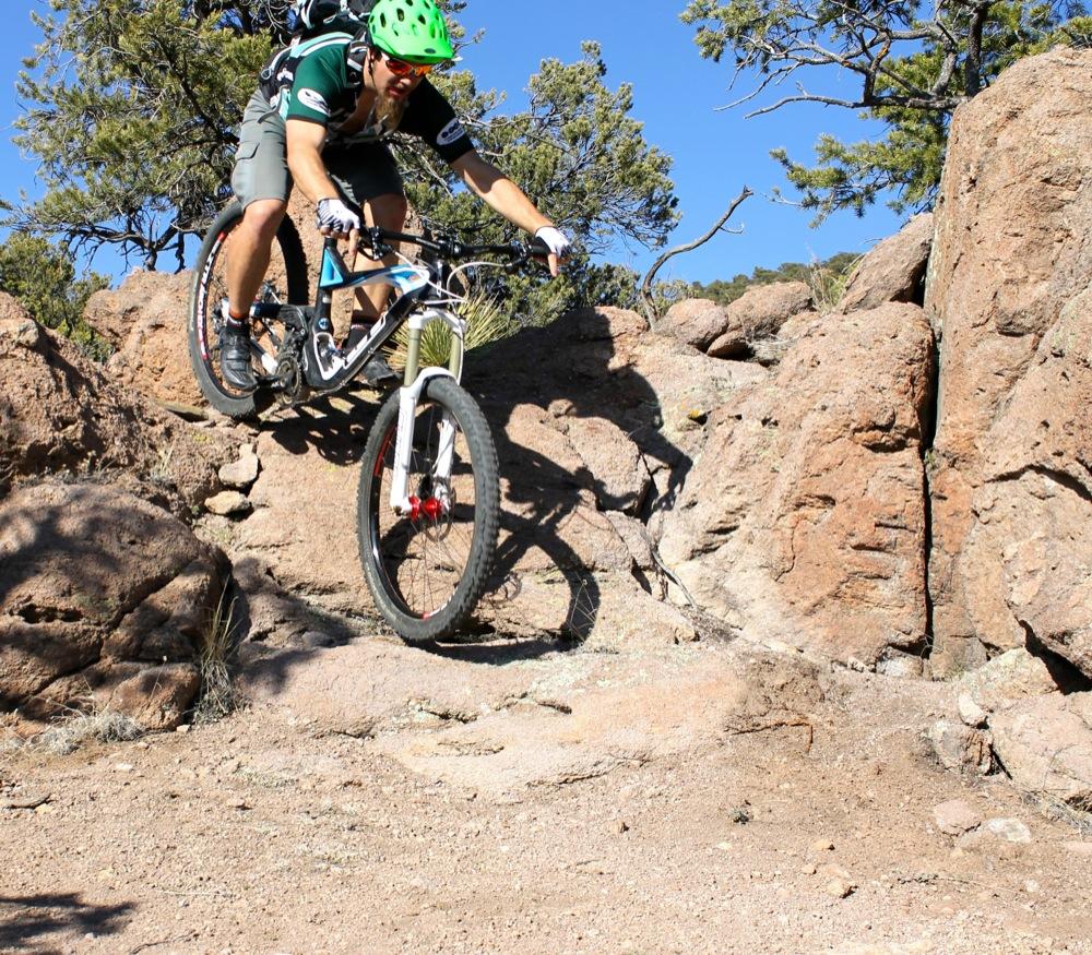 A mountain biker in a green helmet and cycling gear jumps over rocks on a rugged terrain, showcasing his skills on a sunny day with trees in the background. Unkle Nazty mountain bike trail.