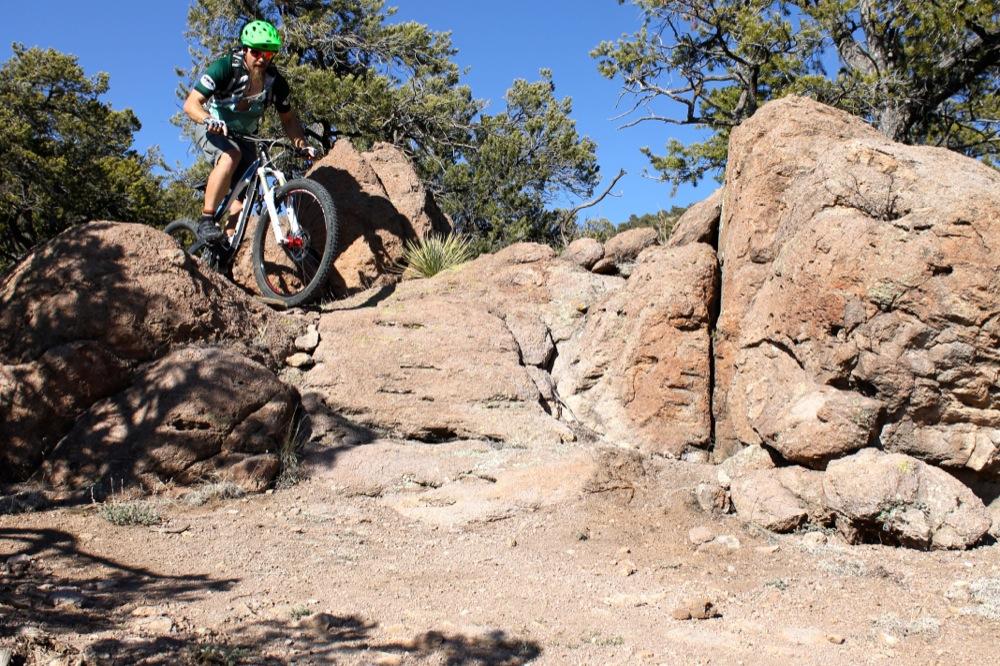 A mountain biker skillfully navigates a rocky terrain, riding over large boulders and surrounded by sparse vegetation under a clear blue sky. Unkle Nazty mountain bike trail.