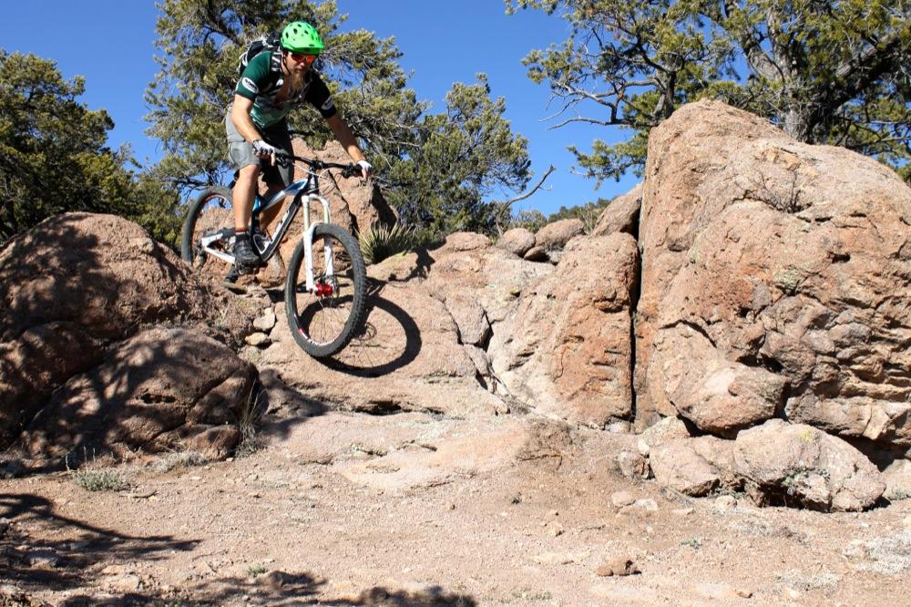 A mountain biker in a green helmet performs a jump over rocky terrain surrounded by trees under a clear blue sky. Unkle Nazty mountain bike trail.