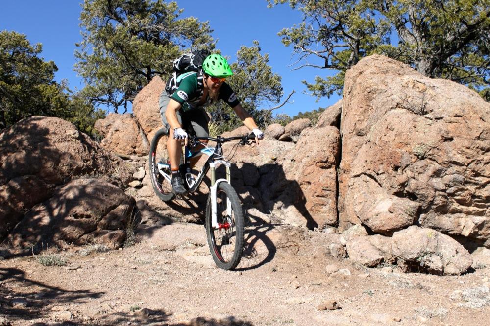 A mountain biker in a green helmet and black backpack jumps over a rocky terrain under a clear blue sky. The rider is wearing shorts and a short-sleeved shirt, demonstrating an action-packed moment in outdoor cycling. Trees can be seen in the background, enhancing the natural setting. Unkle Nazty mountain bike trail.