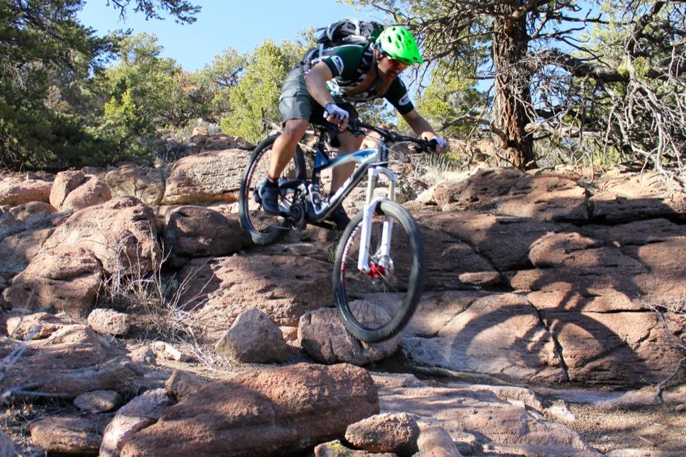 A mountain biker with a green helmet navigates a rocky trail, airborne over a large boulder, surrounded by trees and natural scenery. The sun illuminates the scene, highlighting the rugged terrain. Unkle Nazty mountain bike trail.