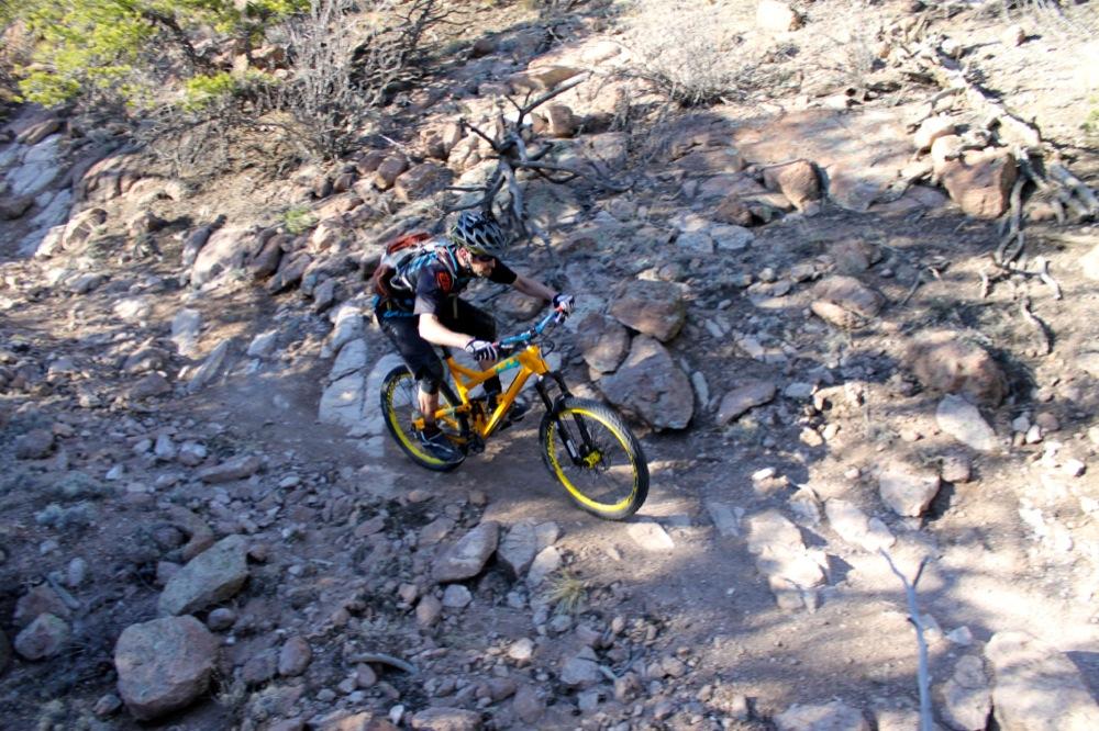 A mountain biker navigates a rocky trail on a yellow bike, wearing a helmet and sports attire. The terrain is rough, with scattered stones and dry vegetation in the surrounding area. Unkle Nazty mountain bike trail.