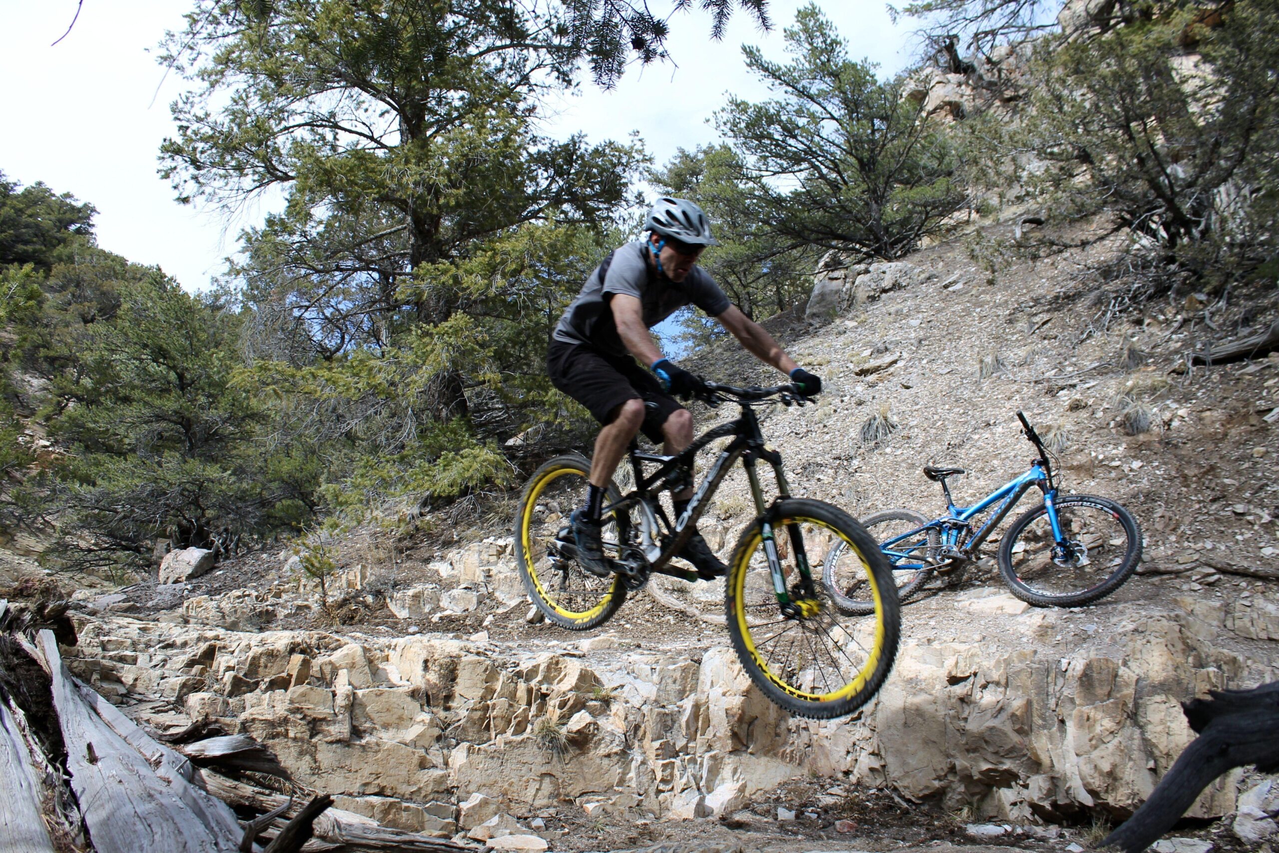 Mountain biker airborne over rocky terrain, with trees in the background and another bike resting on the ground nearby. Cottonwood mountain bike trail.