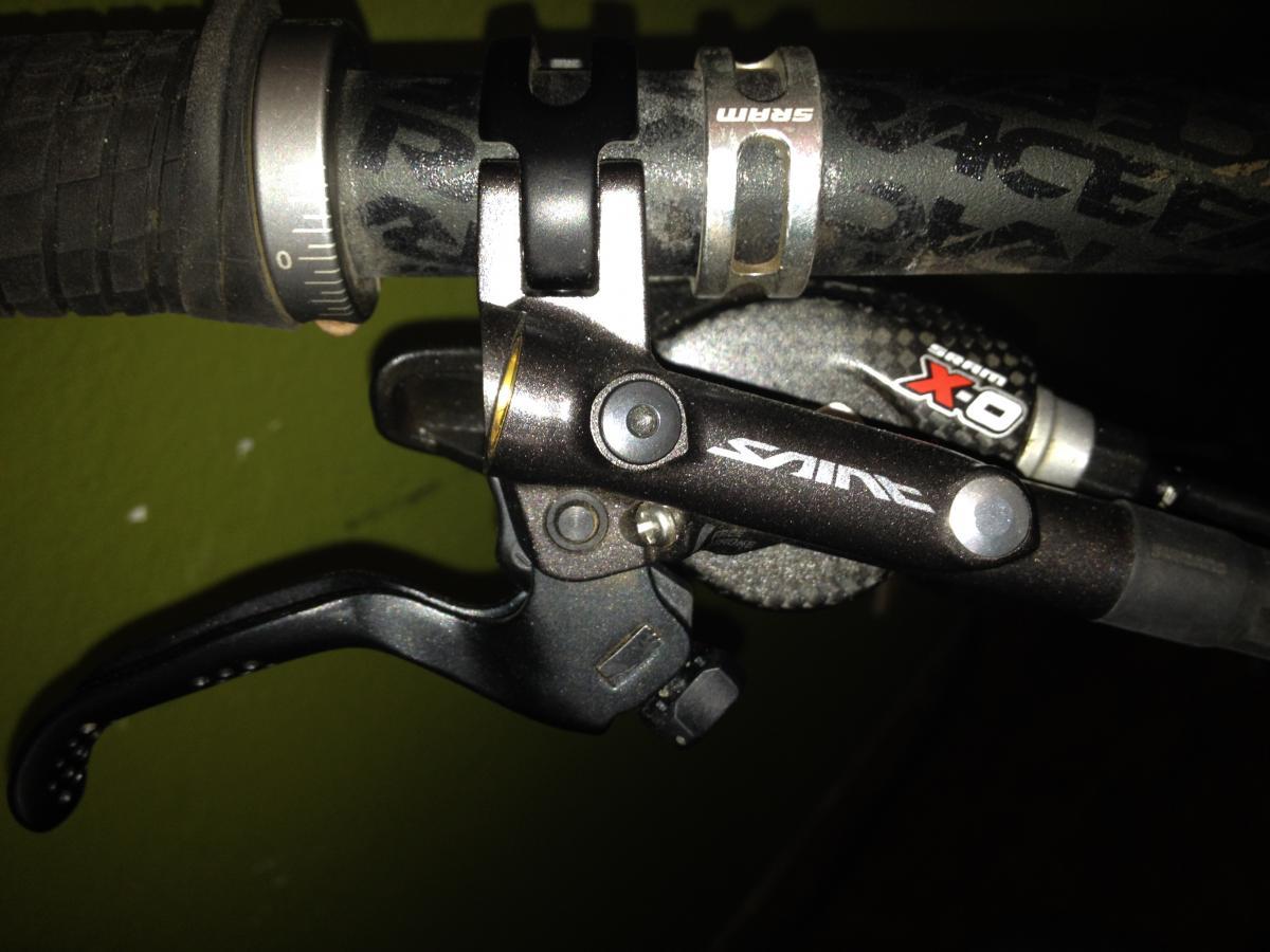 Turner Sultan: Close-up image of a bicycle handlebar featuring a Shimano Saint brake lever. The grip is textured and black, with a grey adjustment ring. A SRAM X0 shifter is also visible, along with part of the handlebar itself. The background is a muted green color.