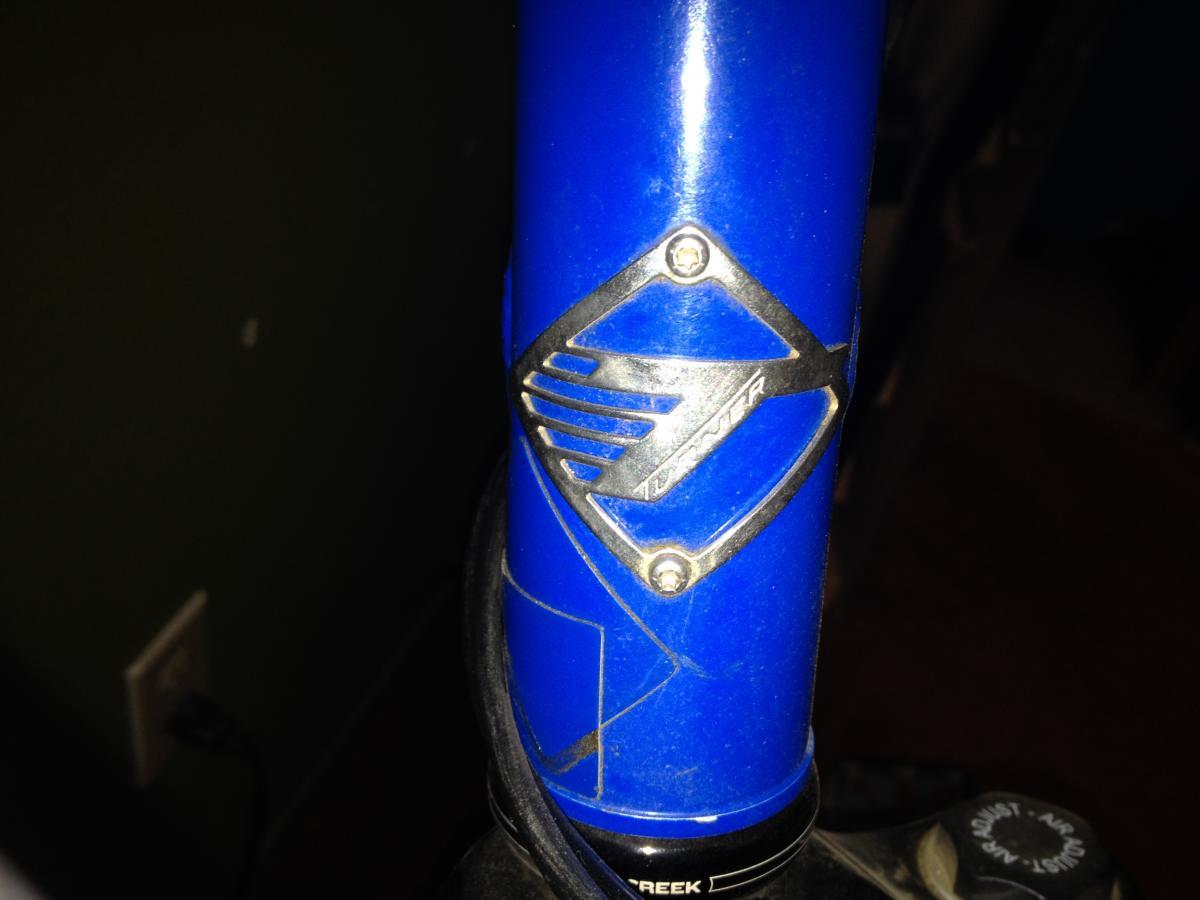 Turner Sultan: Close-up image of a blue bicycle frame with a silver logo emblem. The emblem features a design with angled lines and a diamond shape, positioned on the neck of the frame. The background is dimly lit, highlighting the frame and logo details.