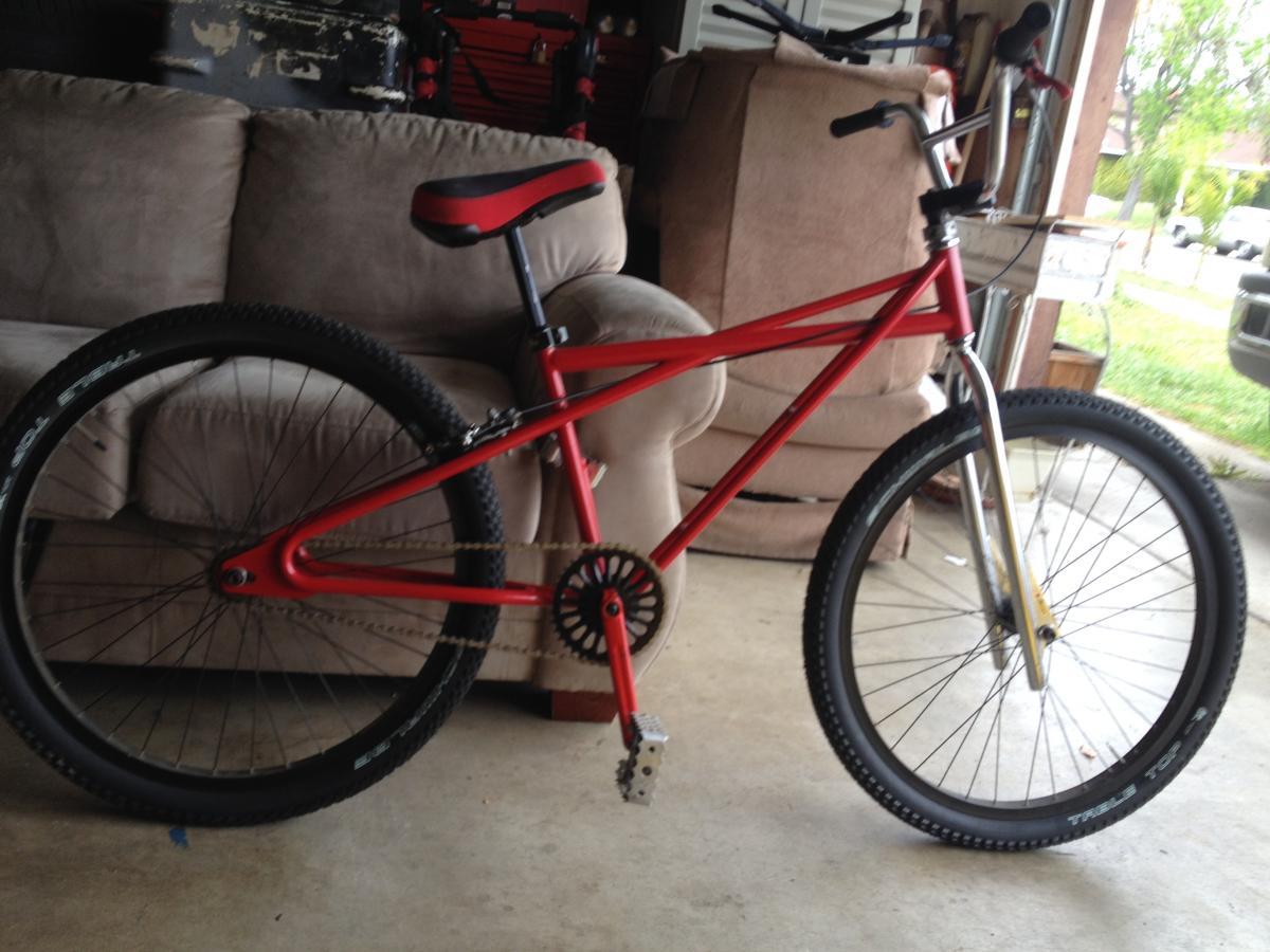 Turner Sultan: A red bike with a sleek frame and large tires, positioned in a garage. There is a beige couch in the background, along with various tools and items visible in the setting. The bike features a single gear and a minimalistic design, suitable for riding in various terrains.