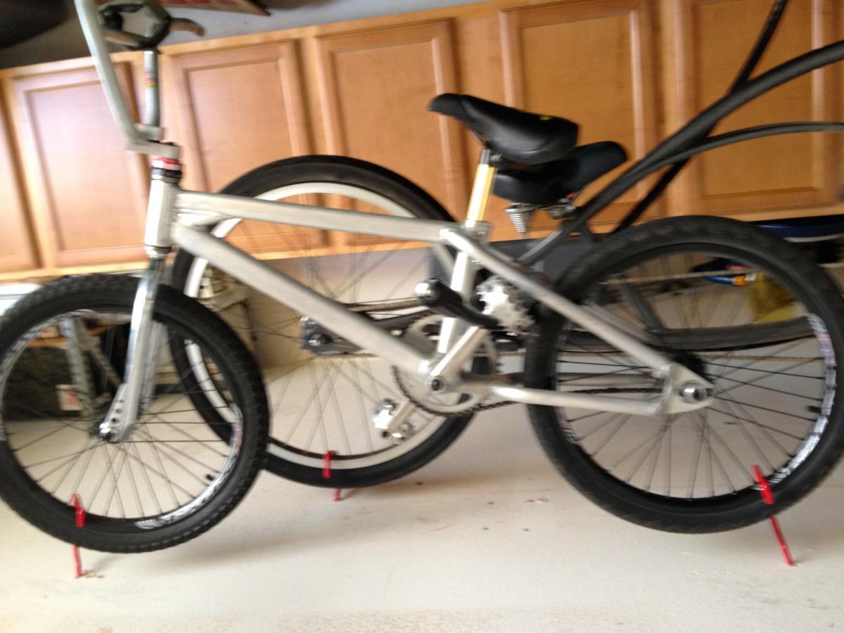Turner Sultan: A silver BMX bike partially leaning against a wall in a garage. The bike features black tires and a black seat, with red wheel stands visible holding the tires in place. The background shows wooden cabinets and some clutter typical of a garage setting.