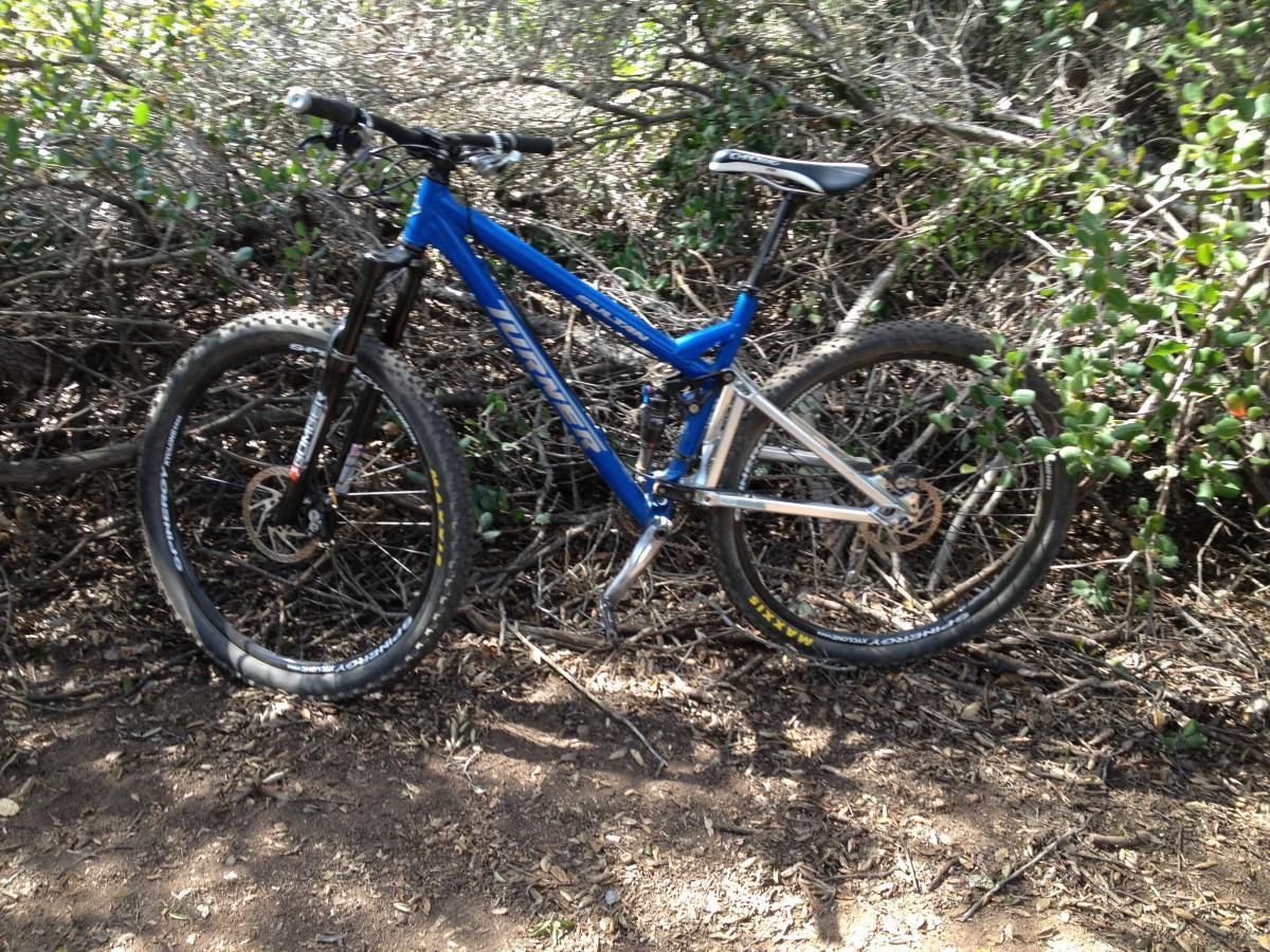 Turner Sultan: A blue mountain bike with silver accents is resting on the ground among bushes and dirt. The bike features thick tires and a sturdy frame, positioned in a natural, outdoor setting.