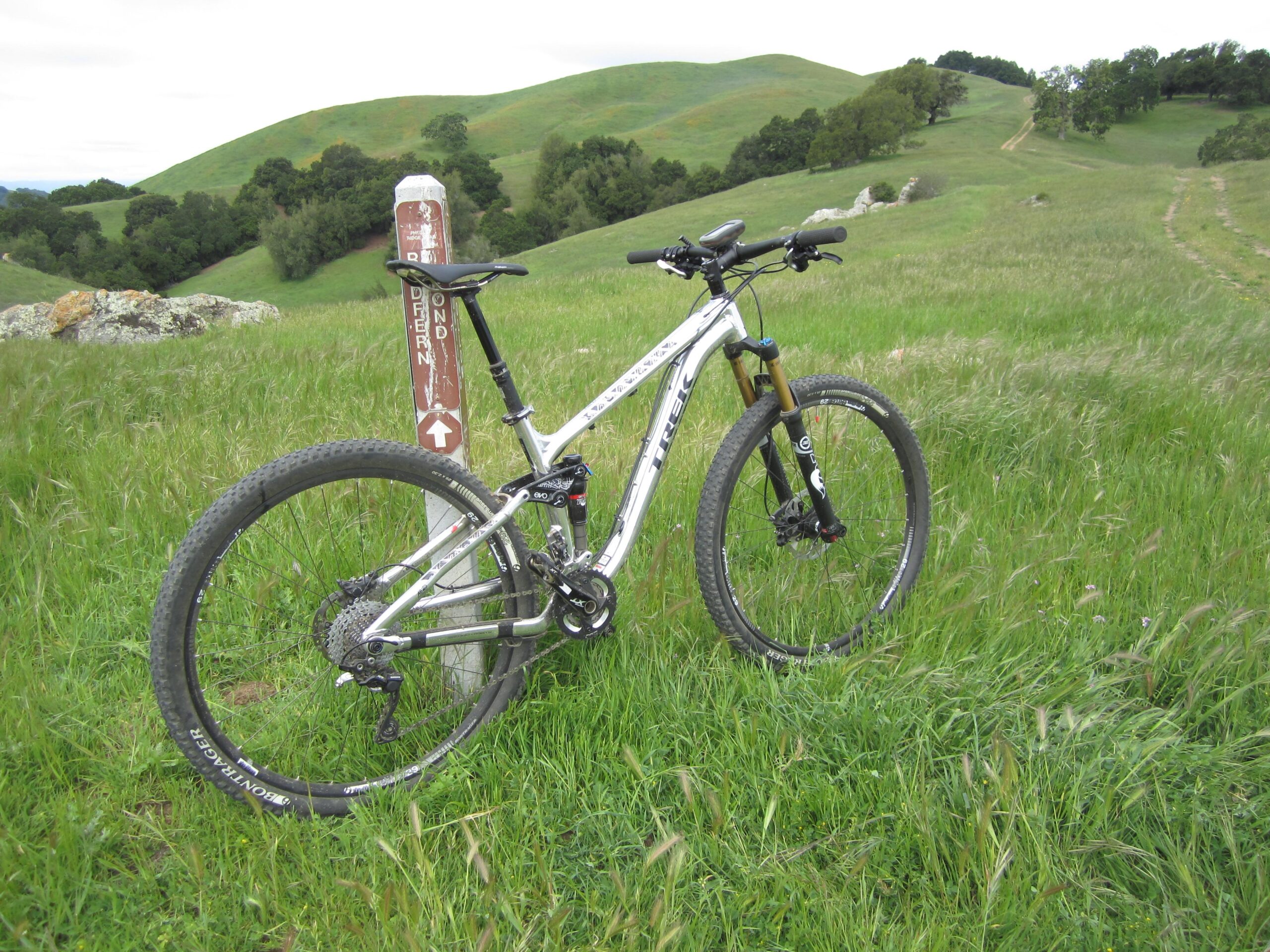 Trek Fuel EX 9: A mountain bike rests against a signpost in a grassy field, with rolling green hills in the background. The scene captures a tranquil outdoor setting ideal for biking and exploring nature.