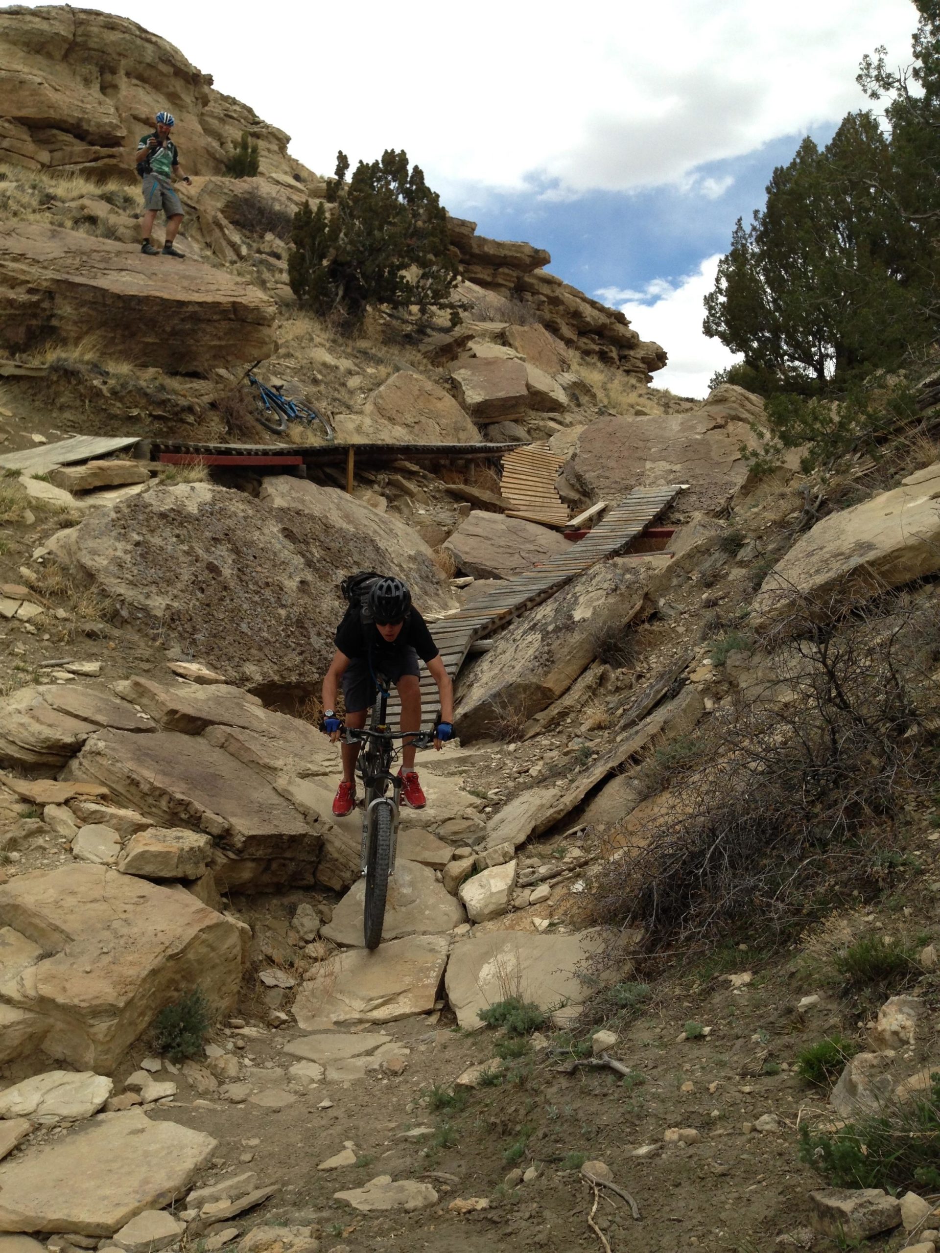 A mountain biker navigating a rocky trail with wooden ramps, surrounded by rugged terrain and sparse vegetation under a partly cloudy sky. Another cyclist is visible in the background, standing on a rock formation. South Shore Lake Pueblo mountain bike trail.