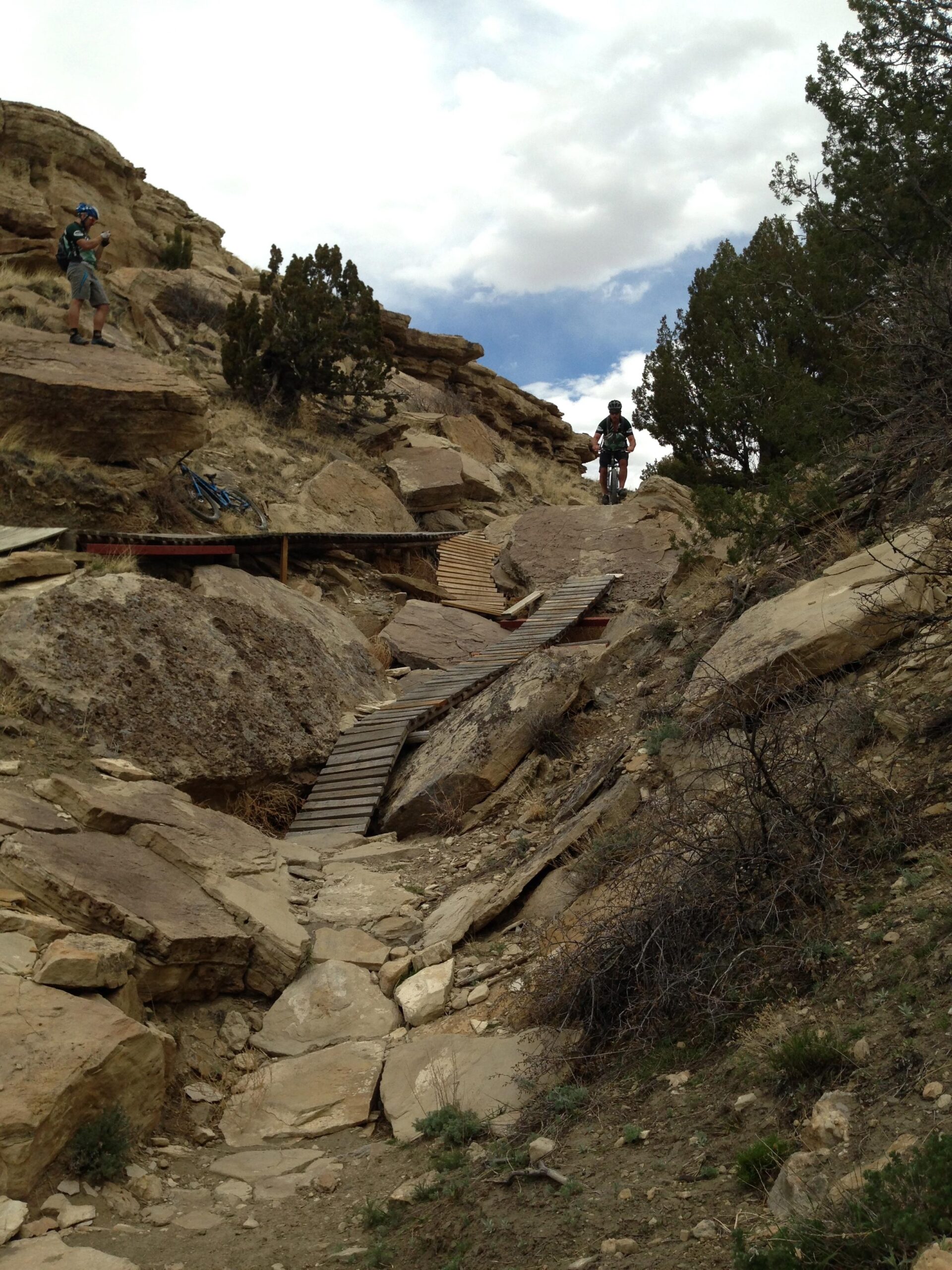 Two mountain bikers navigating a rocky trail with wooden planks in a rugged, outdoor landscape. One rider is ascending the trail while another stands nearby, preparing to take a photo. The scene features rocky terrain, scattered vegetation, and a cloudy sky in the background. South Shore Lake Pueblo mountain bike trail.