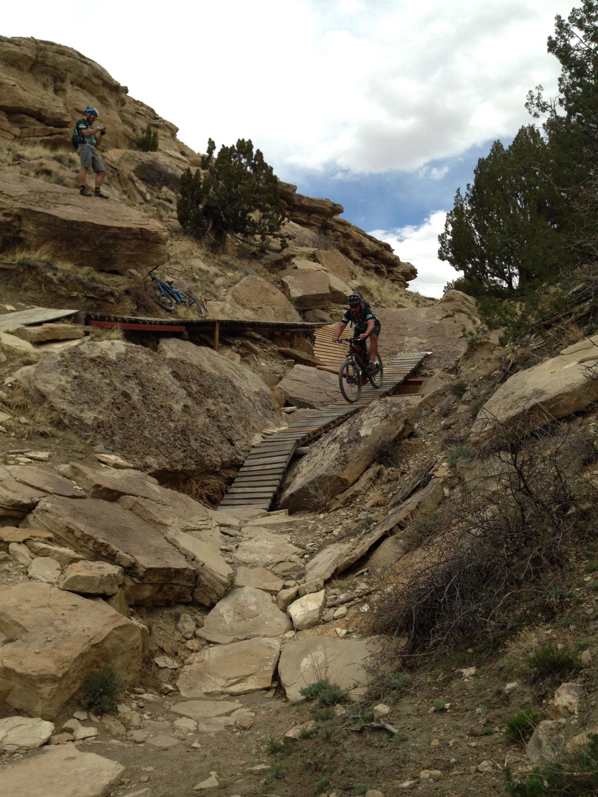 A mountain biker descends a rocky trail, navigating a wooden bridge with steep terrain on either side. A second person stands above, possibly photographing the action, while a bike leans against the rocks nearby. The landscape consists of rugged rocks and sparse vegetation under a partly cloudy sky. South Shore Lake Pueblo mountain bike trail.